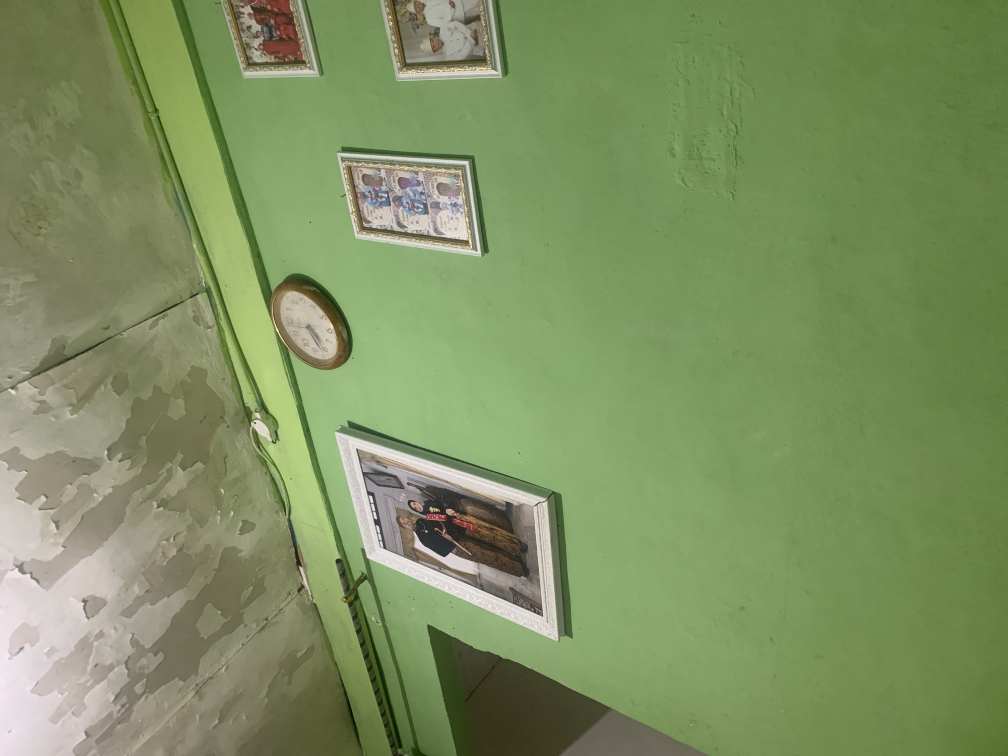 The image depicts a green-painted wall in what appears to be an indoor setting. The ceiling above shows signs of peeling paint. Several framed photographs are displayed on the wall.
On the left, a large framed photograph shows a couple dressed in traditional attire, possibly for a wedding or formal occasion. The man wears a dark jacket over a patterned sarong, and the woman wears a patterned sarong with a dark top and headscarf.
To the right of this large photograph, a circular analog clock with a wooden frame is mounted on the wall.
Further to the right, there are three smaller framed photographs arranged vertically. The top frame shows a group of people, possibly children, in red attire. The middle frame contains multiple smaller images, some featuring cartoon characters. The bottom frame also displays several smaller photographs.
To the far right, another framed photograph shows two individuals in white attire, possibly religious or ceremonial clothing.
The overall impression is that of a residential interior, possibly decorated with personal mementos. Without further context or identifiable landmarks within the image, it is impossible to definitively pinpoint the location in the city of Palembang, Indonesia. The style of the photographs and attire *might* suggest a local cultural context, but this is speculative.