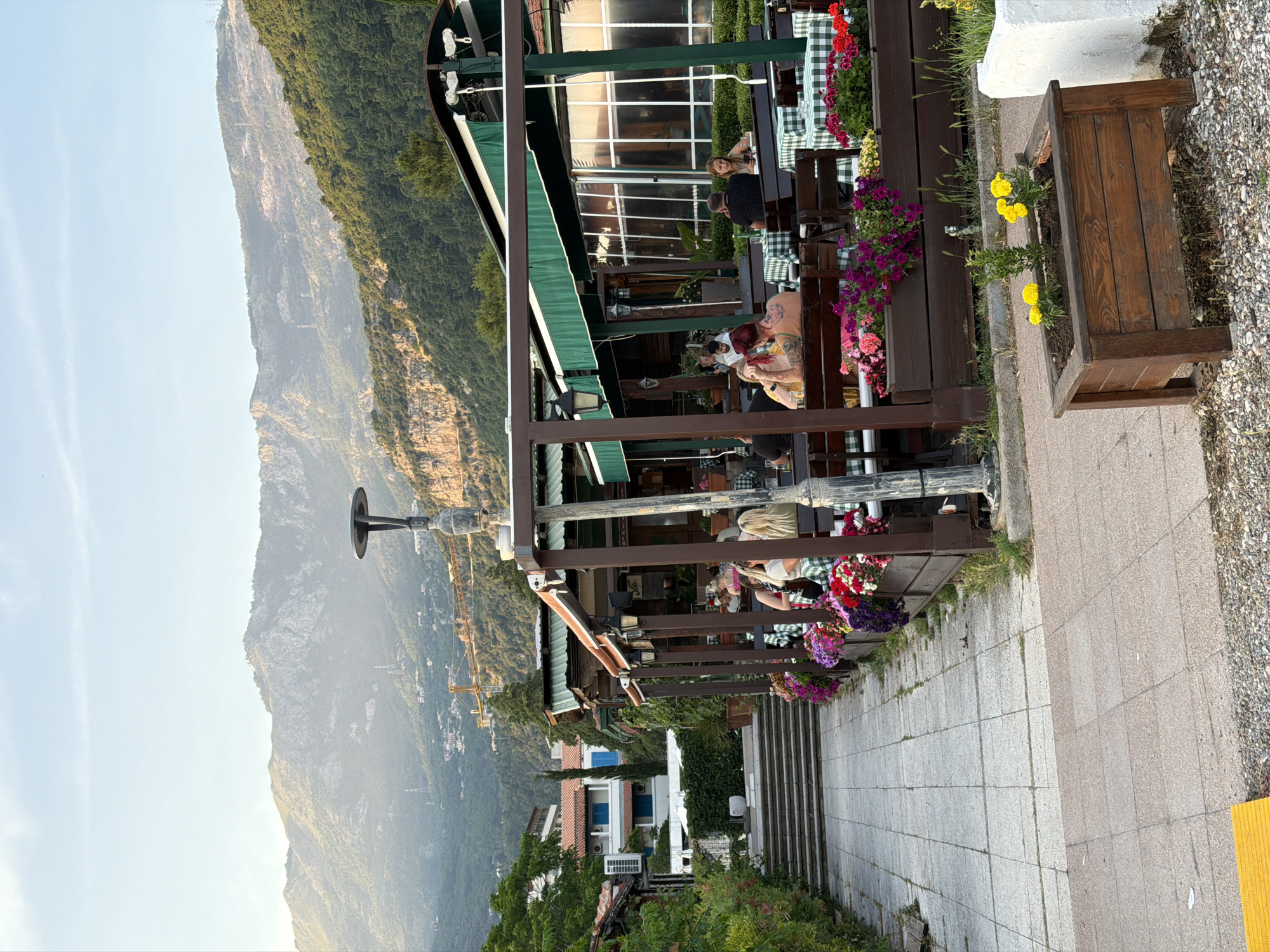 The image shows an outdoor restaurant in Budva, Montenegro, with patrons seated at tables under a green awning. Checkered tablecloths cover the tables. The restaurant has flowers in planters along the edges. A pathway made of paving stones runs alongside the restaurant, and a planter box with yellow flowers is near the pathway. In the background, there is a steep mountain covered in vegetation. Further down, there are buildings with red roofs. A white building with blue details is visible near the bottom. The sky is bright and clear. A street lamp is visible.