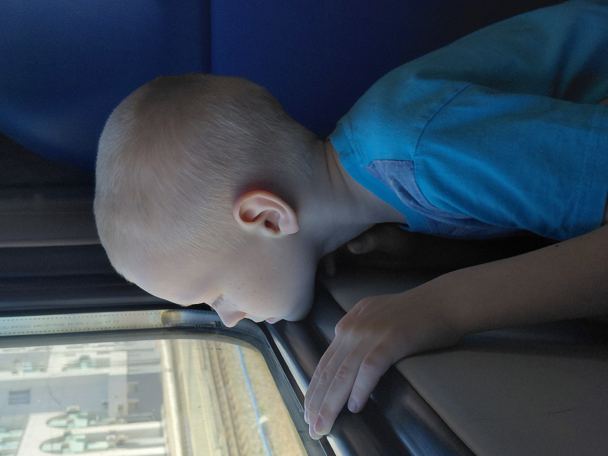 A young boy with short blonde hair and freckles looks out a train window. He is wearing a bright blue t-shirt. His left hand rests on the window frame. The boy is seated on a blue train seat, and the view outside the window appears to be of a city with modern buildings and train tracks. The specific location in Novosibirsk, Russia, cannot be determined from the image.