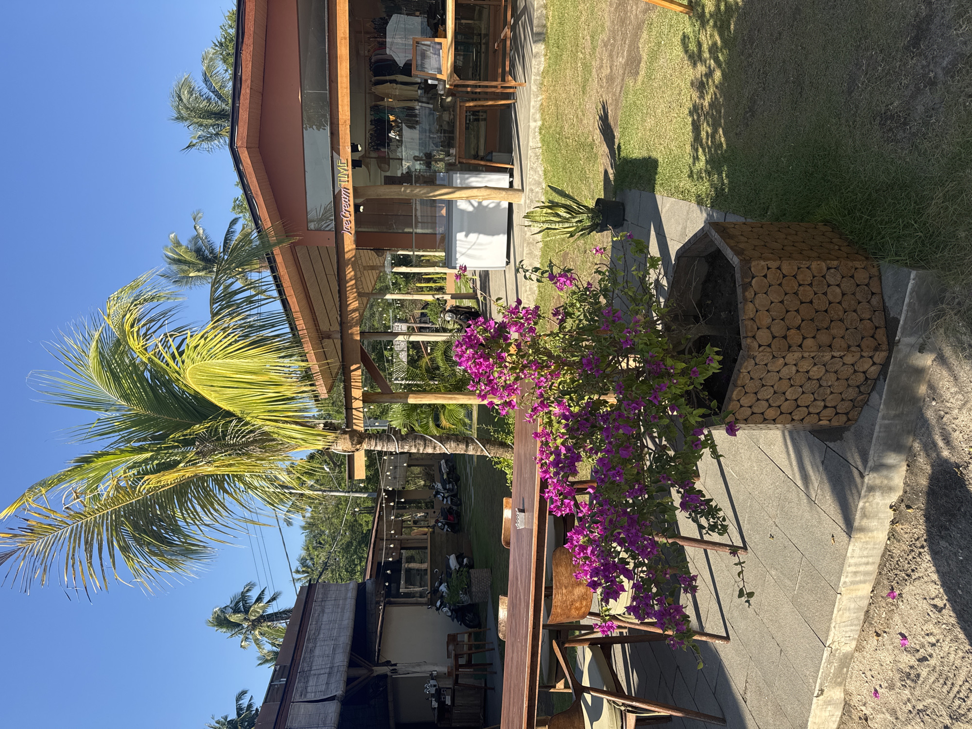 This image captures an outdoor commercial establishment located in Nusa Penida, Indonesia, under a clear, bright blue sky with direct sunlight.
The scene is dominated by two primary buildings and an extensive outdoor area. On the right, a modern single-story building features a reddish-brown exterior, a wide overhanging wooden roof supported by beams, and large glass windows. A yellow sign above the main entrance clearly displays "Ice Cream TIME." Inside, a white commercial freezer unit is visible, along with wooden tables and racks displaying what appears to be merchandise.
To the left, a more rustic, open-air structure with a natural-material roof and rolled-down bamboo blinds provides additional shaded seating. Further in the background, additional low-rise buildings are visible, connected by overhead string lights.
In the mid-ground, a long, dark wooden rectangular table is situated on a paved patio of light grey stone slabs, accompanied by several matching wooden chairs. A small metal ashtray or similar item rests on the table. Directly in the foreground, a vibrant purple bougainvillea bush, rich with blossoms, is contained within a distinctive hexagonal planter constructed from stacked, circular wooden logs. Scattered purple petals are visible on the paved ground and the adjacent sandy soil. Near the "Ice Cream TIME" building, a potted Sansevieria (snake plant) is placed on the grass.
The ground cover alternates between light grey stone paving, areas of green grass, and patches of sandy or dirt ground, particularly in the immediate foreground. In the mid-ground, behind the main table and in front of the background structures, approximately five to six scooters or motorbikes are parked on the grassy area. A large palm tree with both green and yellowed fronds stands prominently in the center-right. No individuals are present or visible within the frame. Strong sunlight casts distinct, elongated shadows from the vegetation and structures across the scene.