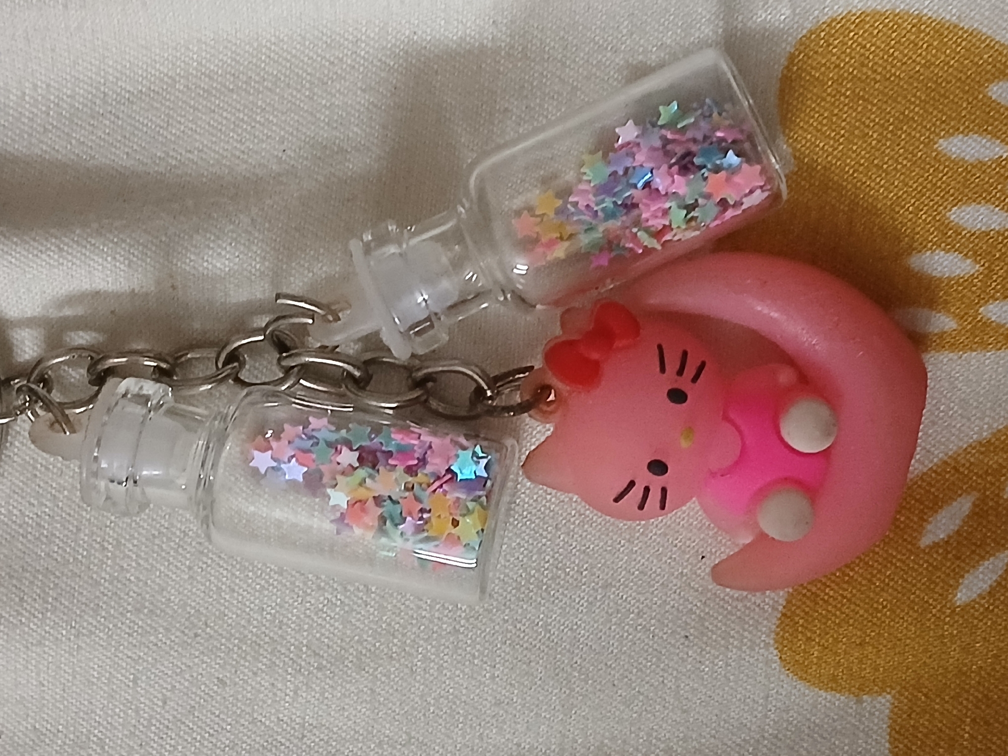 A close-up shot depicts a keyring or keychain lying on a textured surface. The keyring consists of a silver chain, two small, clear plastic bottle-shaped containers filled with colorful star-shaped confetti, and a pink, stylized Hello Kitty figure. The Hello Kitty figure has a red bow, black whiskers, and white "feet." The textured surface appears to be fabric, with a white and yellow patterned area visible on the right. There is no information available in the image that would indicate that the location is in Bengaluru, India.