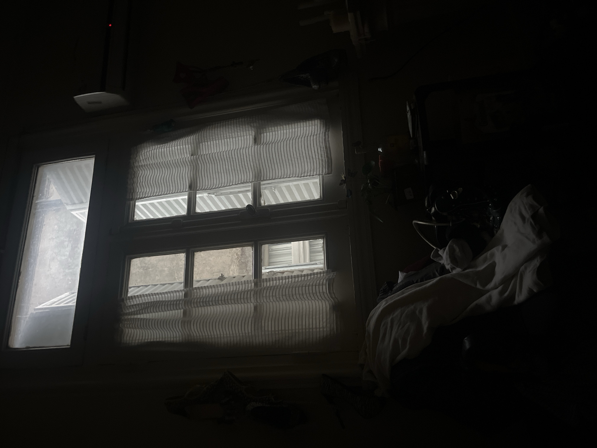 The image depicts a dimly lit room, possibly in Buenos Aires, Argentina. Two windows are visible, one with clear glass and the other covered with a striped curtain. Outside, the cityscape is visible through the uncovered portion of the window. An air conditioning unit is mounted above the window. A bed with white sheets is located on the right side of the frame, obscured by darkness. Unidentifiable objects are scattered on the floor in the foreground. The room is dark, illuminated primarily by the light coming through the windows.