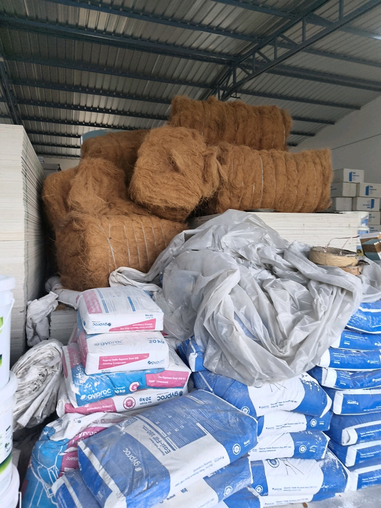 The image depicts a warehouse interior in Coimbatore, India, filled with construction materials. Prominently displayed are several large bales of coir fiber, likely for insulation or packaging, stacked in the center and to the right. To the left, stacks of what appear to be drywall or gypsum boards are visible. In the foreground and mid-ground, numerous bags of gypsum compound, such as "Gyproc" jointing compound and ready-mix plaster, are neatly stacked. Some of these bags are covered with white plastic sheeting, suggesting protection from dust or moisture. The environment appears to be a storage facility with a corrugated metal roof and industrial lighting, indicative of a commercial or industrial setting within Coimbatore. No people are visible in the image.