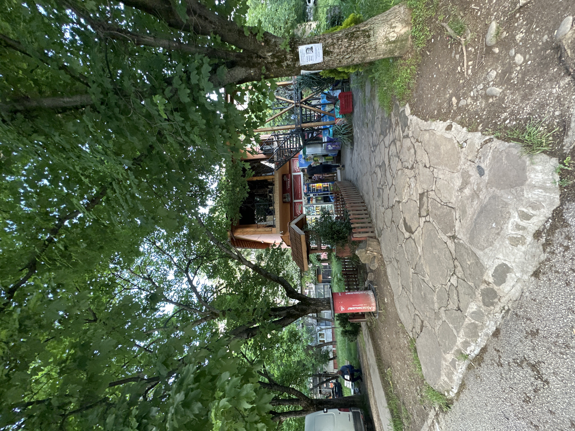 The scene is an exterior view of a wooden structure, likely a small shop or restaurant, situated in Lovech, Bulgaria. The building has a light brown facade and a multi-level layout with a staircase leading to an upper level. A variety of items, possibly merchandise, are displayed inside. A bench and decorative plants adorn the space in front of the building.
The location is surrounded by greenery, including large trees with full foliage. The ground is a mix of stone pathways and grassy areas. A sign is affixed to one of the trees. A red advertising stand is near the building. Further in the background, there is a van and a man working in the vicinity.