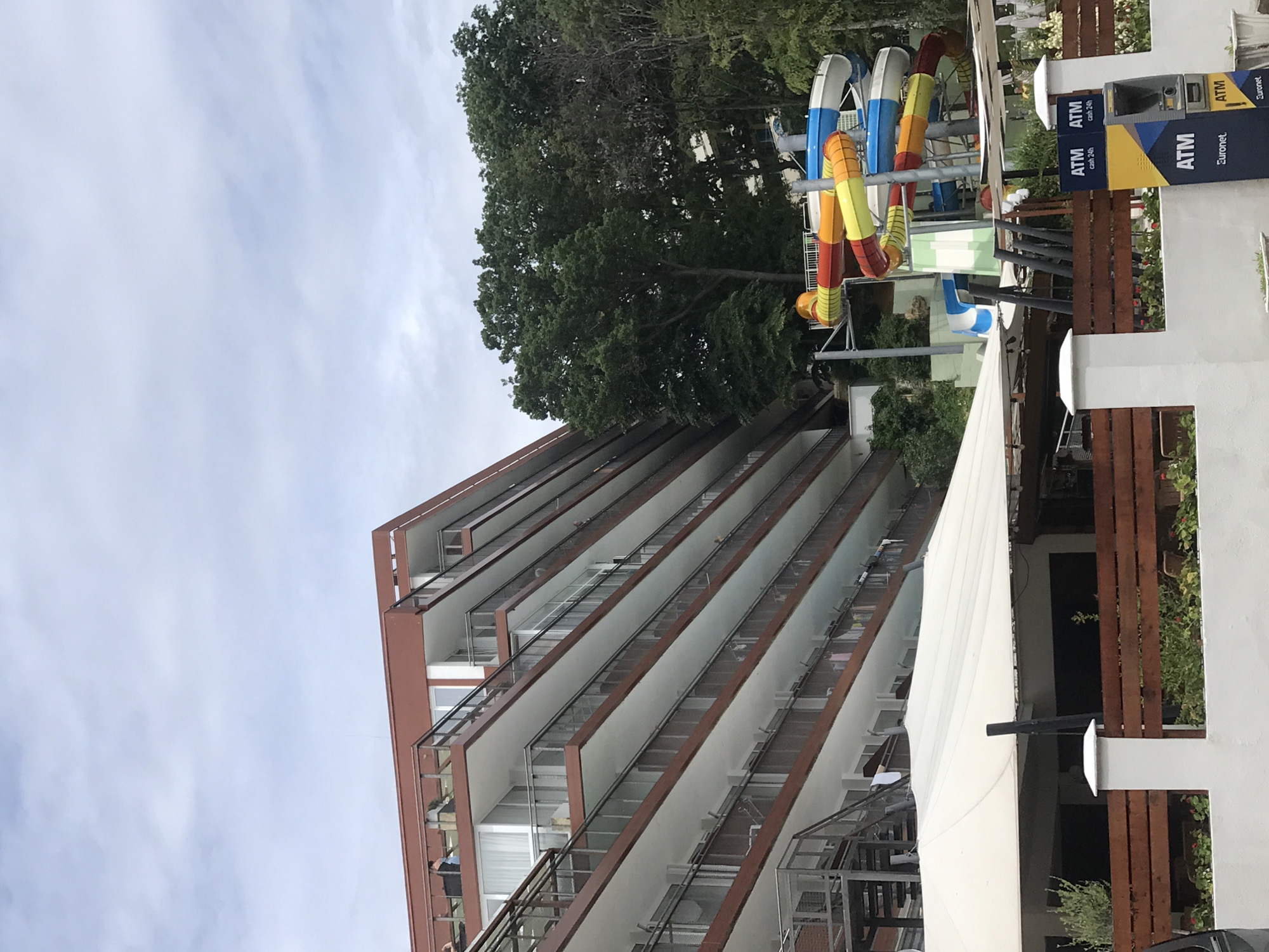 The image shows a large, multi-story hotel with a red-brown exterior and multiple balconies, likely located in Varna, Bulgaria. To the right of the hotel, a colorful water slide complex is visible. In the foreground, a white canopy covers an outdoor area, and a white fence with wooden accents separates the area from the street. An ATM machine is also visible in the foreground. The sky is overcast with clouds.