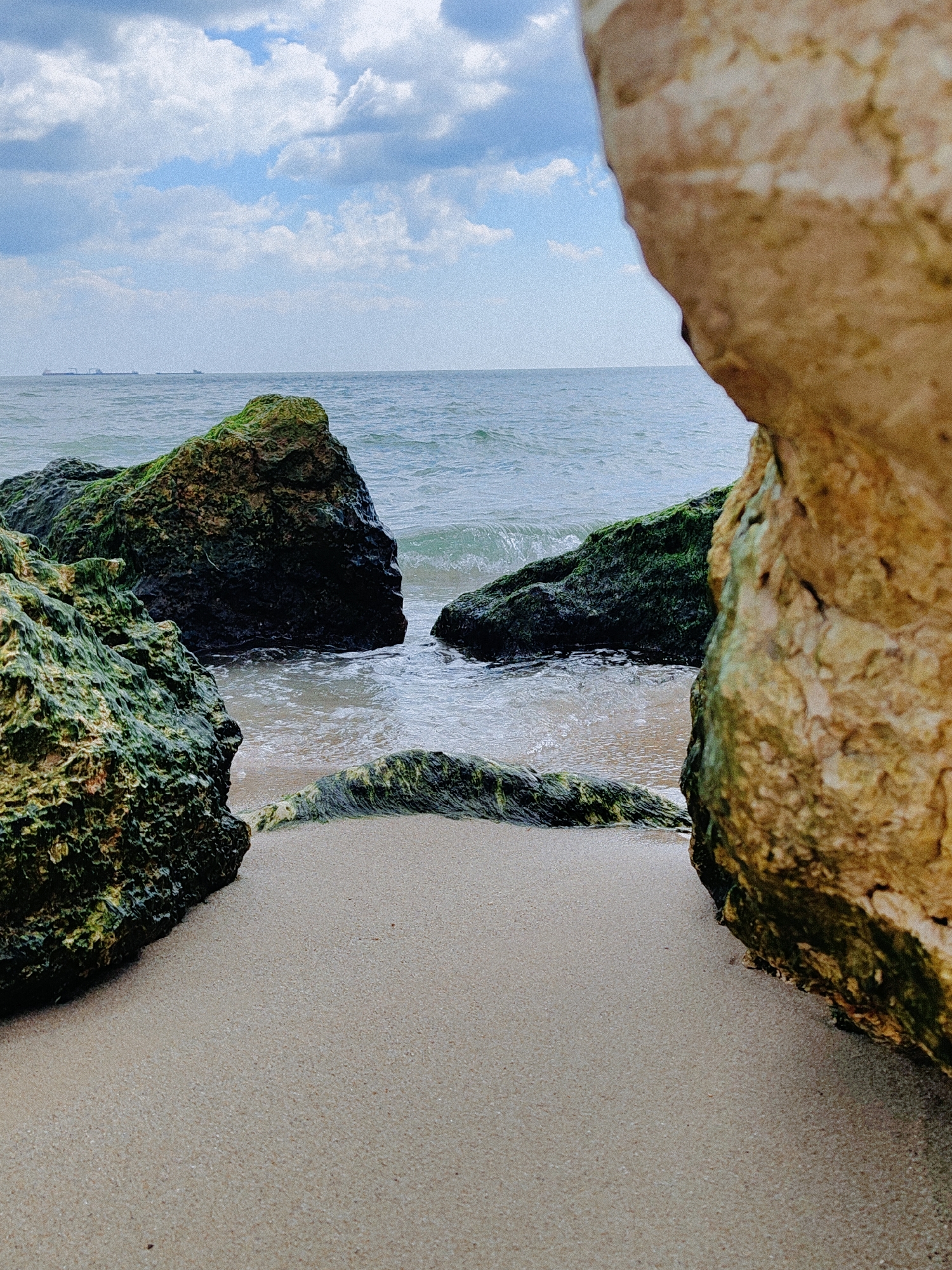The image shows a beach scene in Varna, Bulgaria. Large rocks covered with green algae frame a view of the sea and sky. Sand fills the foreground between the rocks. Waves are gently lapping at the rocks, and several ships can be seen on the horizon. The sky is partially cloudy, with blue patches visible.