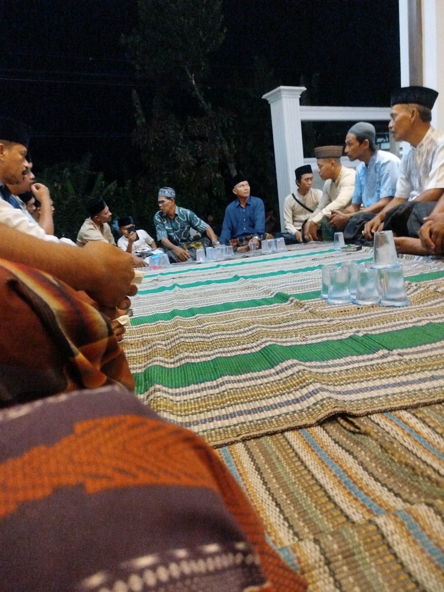 A group of men are gathered outdoors at night, seated on a woven mat in what appears to be a residential area of Burneh, Indonesia. The lighting suggests it is evening or night. The men are dressed in traditional Islamic attire, including peci caps and modest clothing. Several of them are wearing long-sleeved shirts and sarongs. Some are engaged in conversation, while others are looking at their phones or simply sitting. Clear plastic cups are arranged on the mat in front of some of the men, some of which appear to be filled with water. In the background, there is foliage and a white pillar, possibly part of a building. The overall atmosphere suggests a casual social gathering or meeting.