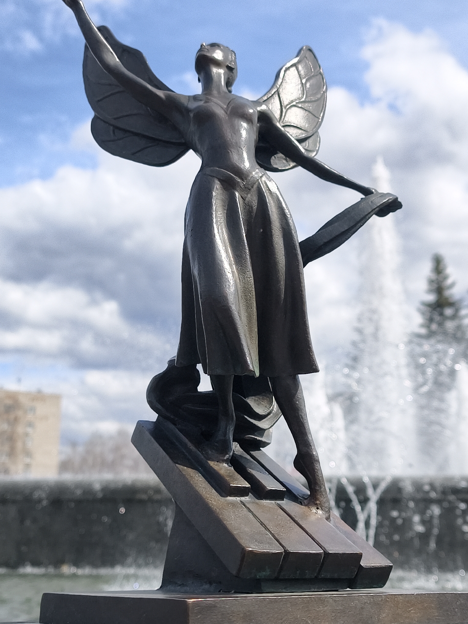 The image captures a bronze statue of a graceful, winged female figure, possibly an angel or fairy, set against a bright, partly cloudy sky. The figure is depicted in a dynamic pose, with her head tilted upwards, eyes closed, and one arm reaching towards the sky. Her other arm extends to the side, holding a flowing piece of fabric or ribbon. She wears a long dress and stands on the keys of a piano, with one foot delicately balancing on the edge of a key, suggesting motion or dance. Her wings are detailed, resembling those of an insect or butterfly.
In the blurred background, a large fountain sprays water high into the air, adding a sense of movement and freshness to the scene. Beyond the fountain, a distant multi-story building and some dark evergreen trees are visible, indicating an urban or park setting. The lighting suggests it is daytime, with sunlight reflecting off the statue's metallic surface, creating bright highlights. The overall scene evokes feelings of aspiration, music, and ethereal beauty.
There is no visible text in the image. The location context provided is Chaikovsky, Russia.