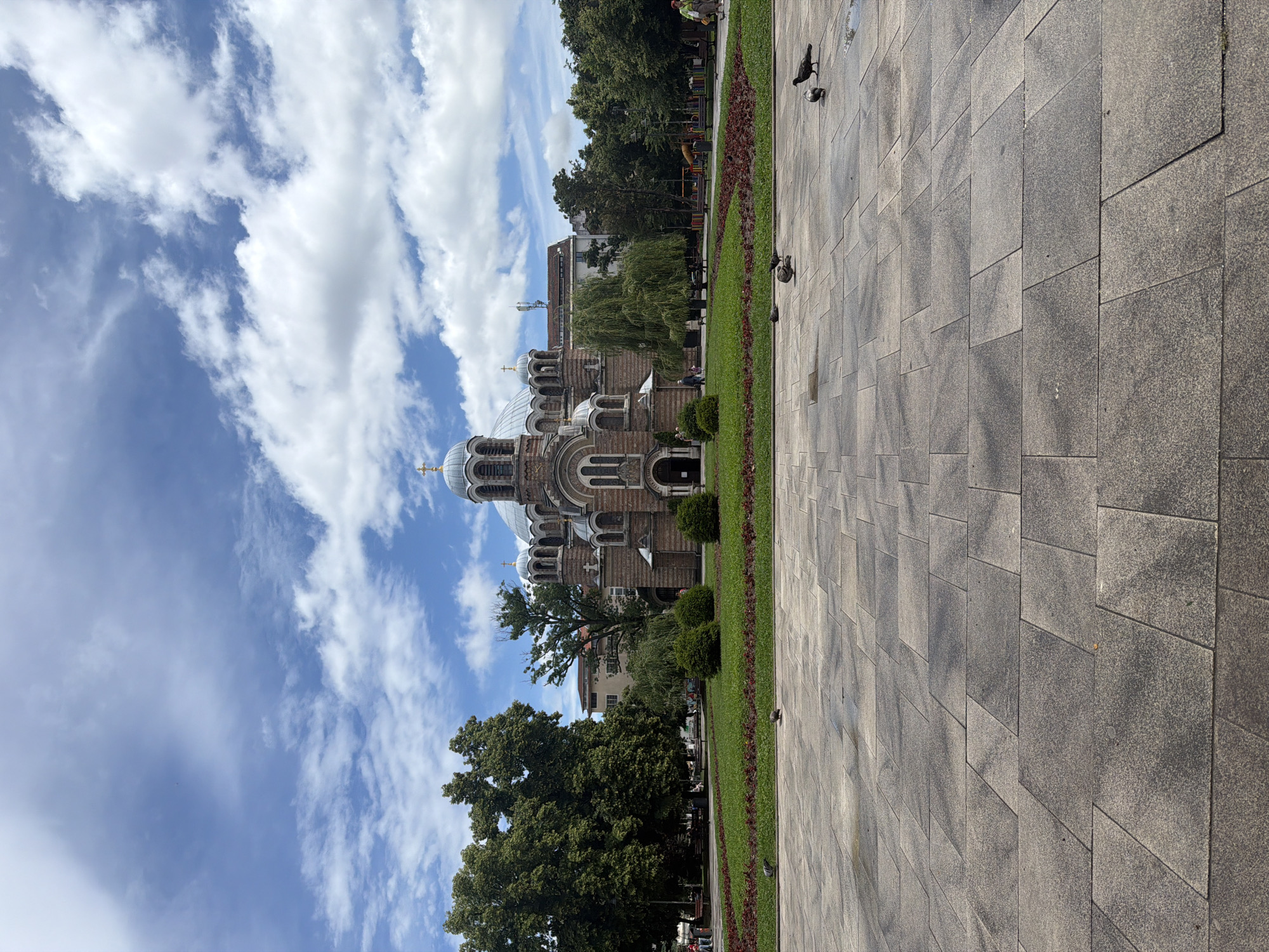 The image displays an outdoor urban scene dominated by a large, ornate Orthodox church on the left side, seen under a bright, partly cloudy blue sky. The church features intricate brickwork and multiple domes, some with light, possibly gilded caps, and others darker, contributing to its grand appearance.
In the foreground and extending towards the right, a wide expanse of paved ground is visible, consisting of light and dark grey rectangular and square paving stones. Several pigeons are scattered across this paved area. A well-maintained strip of green grass, interspersed with reddish-purple flowering plants, horizontally traverses the mid-ground, creating a visual divide between the paved area and the church and surrounding greenery.
Behind the grass strip, numerous mature trees with lush green foliage stand, suggesting a park or landscaped garden adjacent to the church. Beyond these trees and the church structure, other urban buildings can be seen in the distance. A few indistinct human figures are visible in the background, walking or resting among the trees and distant structures.
The weather appears clear and sunny, with scattered white cumulus clouds across the blue sky, indicating a pleasant day. The shadows cast on the paving stones suggest it is mid-day or early afternoon. The architecture of the church and the overall setting align with typical urban landscapes in Sofia, Bulgaria, consistent with the St. Nedelya Church. No visible text is present in the image.