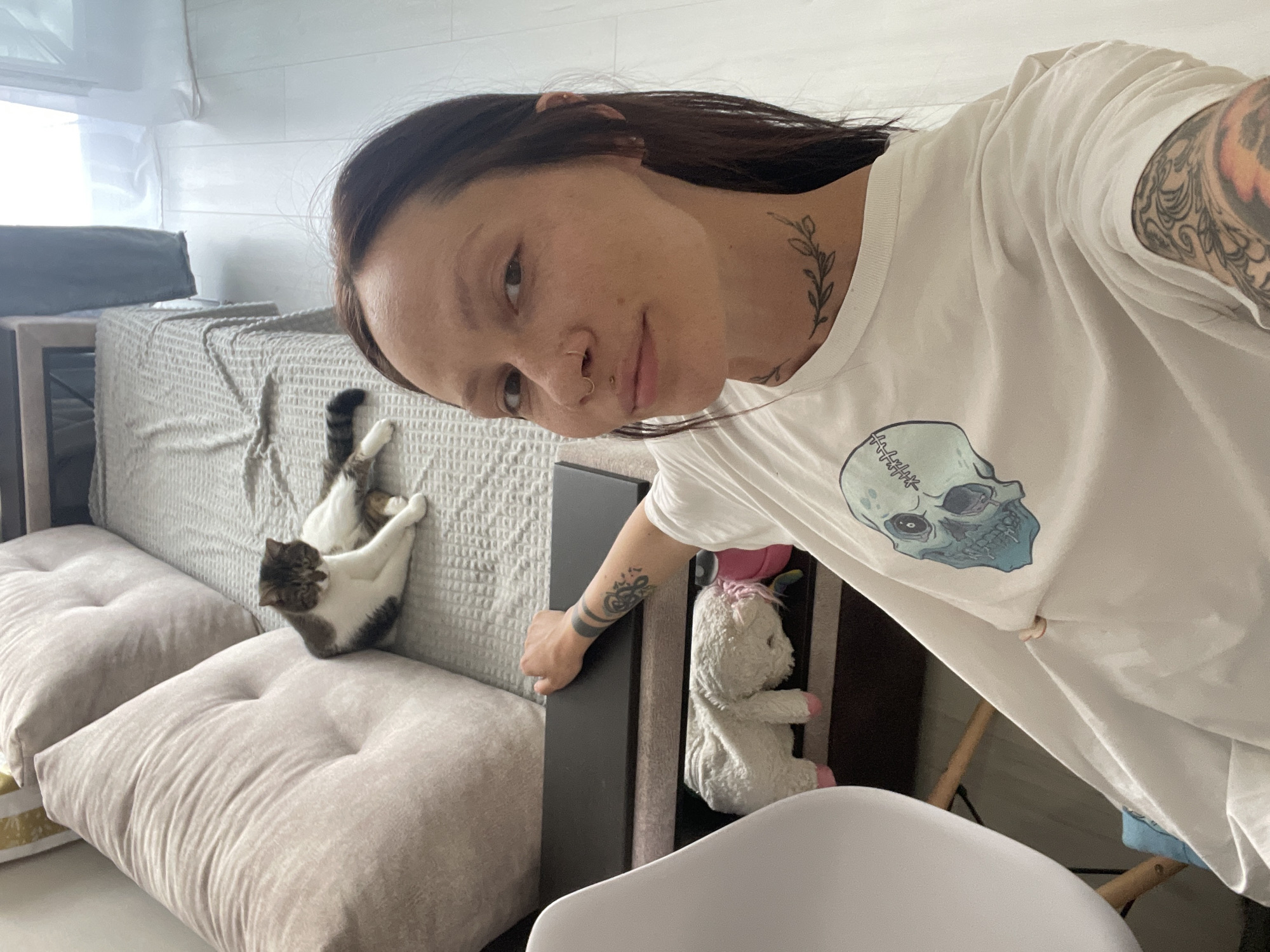 A woman with dark hair, tattoos on her neck and arm, and facial piercings is taking a selfie in what appears to be an indoor setting. She's wearing a white t-shirt with a skull graphic. Behind her is a sofa with light-colored cushions and a patterned throw. A cat is lying on the sofa in a relaxed position. Beside the sofa is a dark-colored piece of furniture with a white stuffed animal tucked inside. A white chair is in the foreground. There is no evidence suggesting this photo was taken in Pervomaysky, Russia.