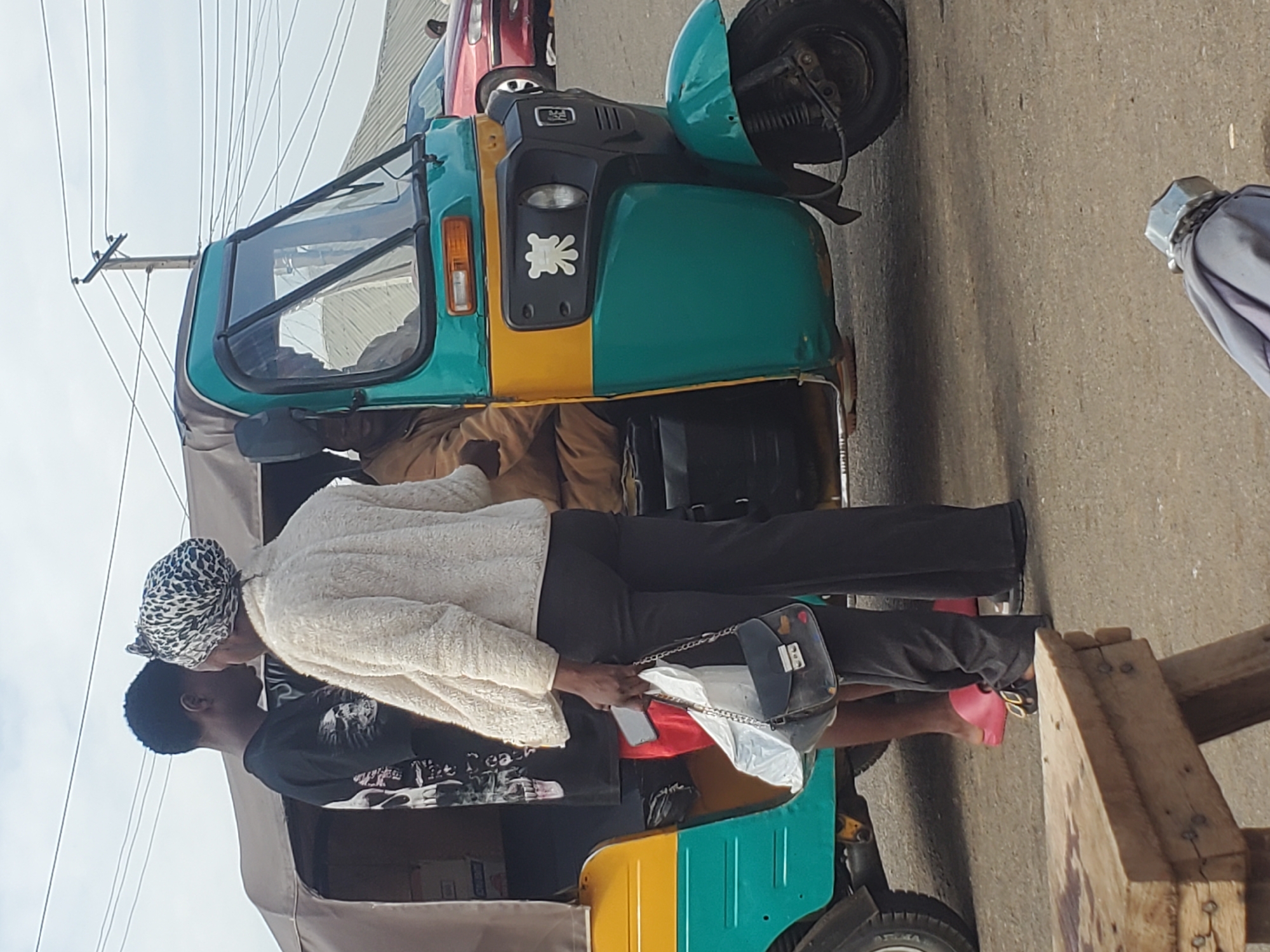 The image shows a scene in the city of Gwong, Nigeria. In the foreground, a wooden table is visible. To the right of the table, a trike is parked, with a person sitting in the driver's seat and two individuals standing next to it. The trike is teal and yellow with a tan canopy. The person on the left is wearing a black t-shirt with a skull design and red shorts. The person on the right is wearing a white fuzzy jacket and black pants, and carrying a black purse and white plastic bag. A red car is visible in the background. The sky is overcast.