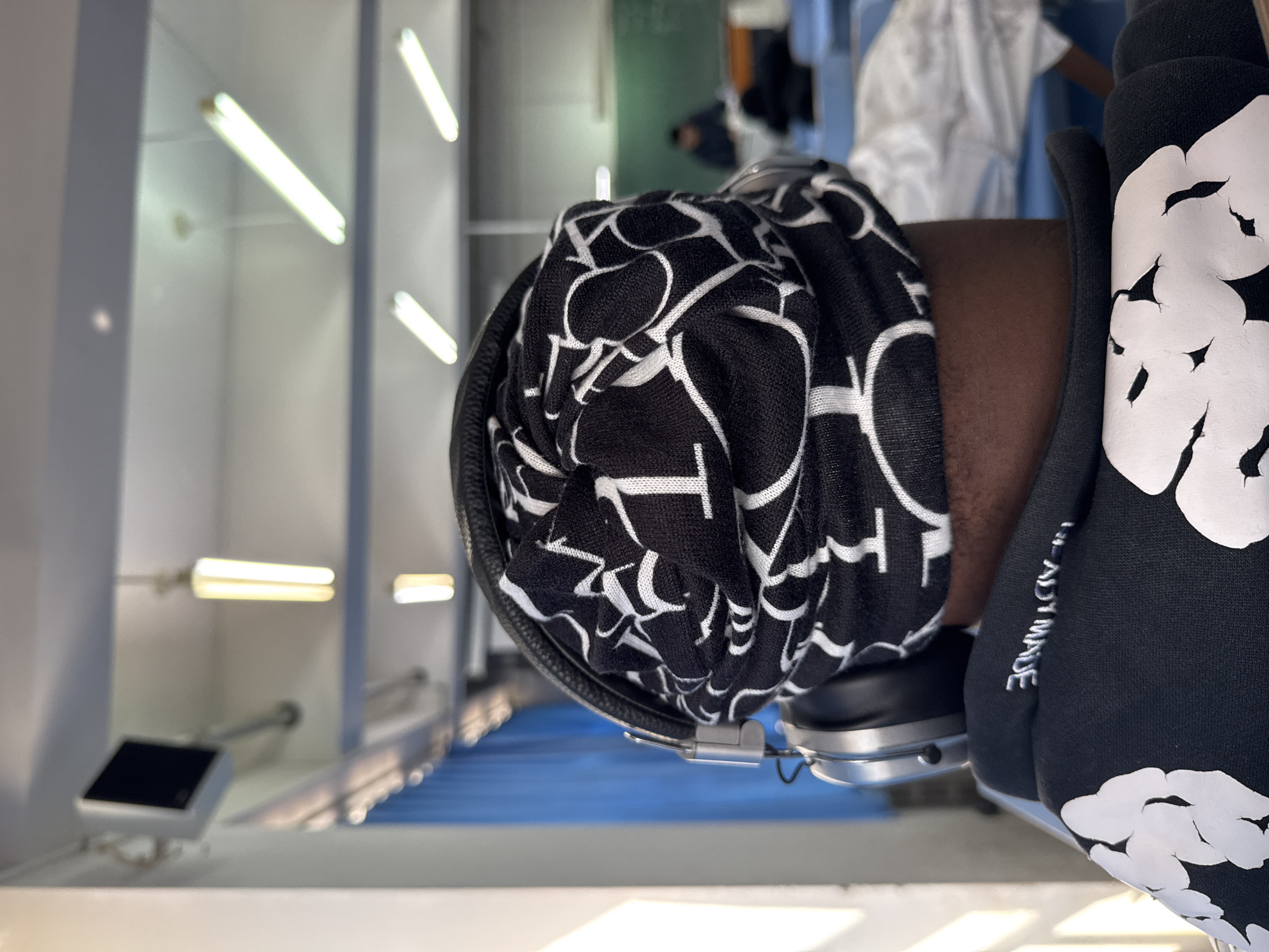 The image presents a close-up, rotated view of a person's upper arm and shoulder in an indoor setting, likely in Zhongtun, China. The person has dark skin and is wearing a black hoodie adorned with large, abstract white designs, resembling distorted clouds or splashes. On the side of the hoodie's sleeve, white embroidered text is partially visible, appearing to spell "Y MADE," which might be part of "READY MADE."
Resting against the person's upper arm is a black and white patterned garment, possibly a scarf or bandana, with intricate white shapes that resemble interconnected letters like 'C' or 'O' against a black background. A pair of silver and black over-ear headphones are positioned under this garment, with one earcup visible.
The background is an indoor environment, characterized by bright, linear fluorescent light fixtures mounted on a light-colored wall or ceiling. A blurred figure in a white top and blue pants is faintly visible further in the background. A prominent blue surface, possibly a floor or a large architectural feature, stretches into the distance. On the lower left, a small, black-screened device on a white stand is visible. The scene suggests a modern public or commercial space.