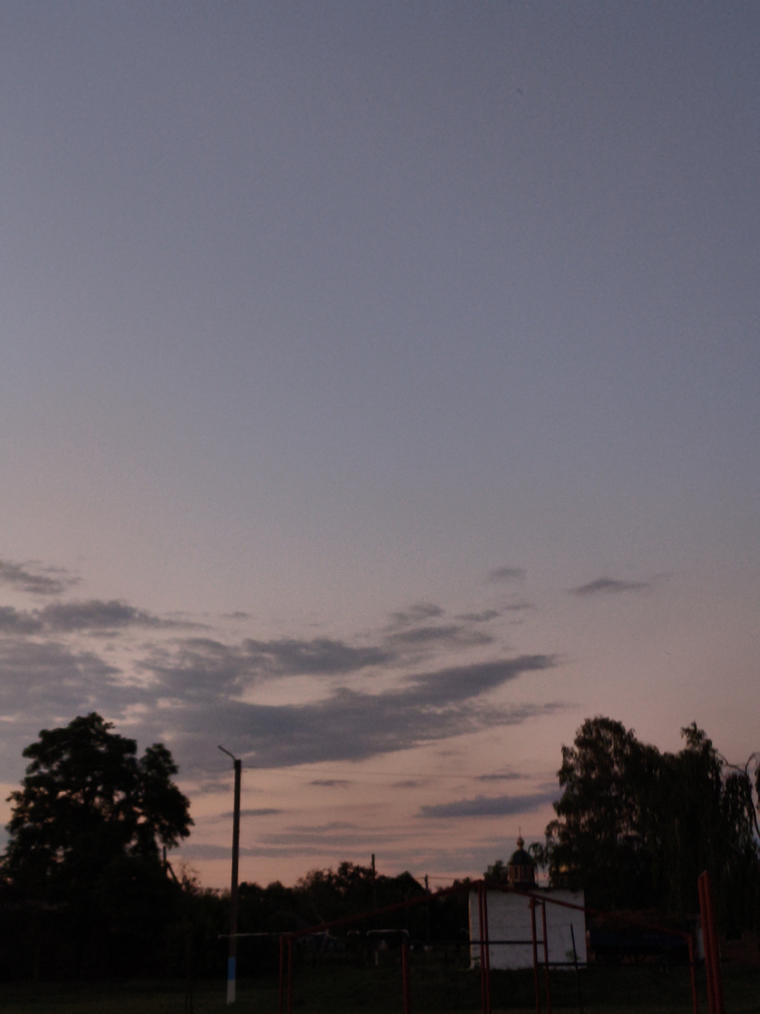 This image captures a rural scene at dusk in Ustyvytsia, Ukraine. The sky is a gradient of pale blue at the top, transitioning to soft pinks and oranges near the horizon, with scattered dark clouds. In the foreground, silhouetted against the fading light, is a large, dense tree on the left side of the frame. To the right of the tree, a utility pole stands vertically, its top extending into the sky. Further to the right, a white building with a dark, domed structure on top, possibly a church or chapel, is visible. The ground is dark and appears to be a grassy area, with indistinct shapes of other trees and structures in the background. The overall atmosphere is peaceful and serene, characteristic of the end of a day in a rural setting. There are no people or animals visible in the image.