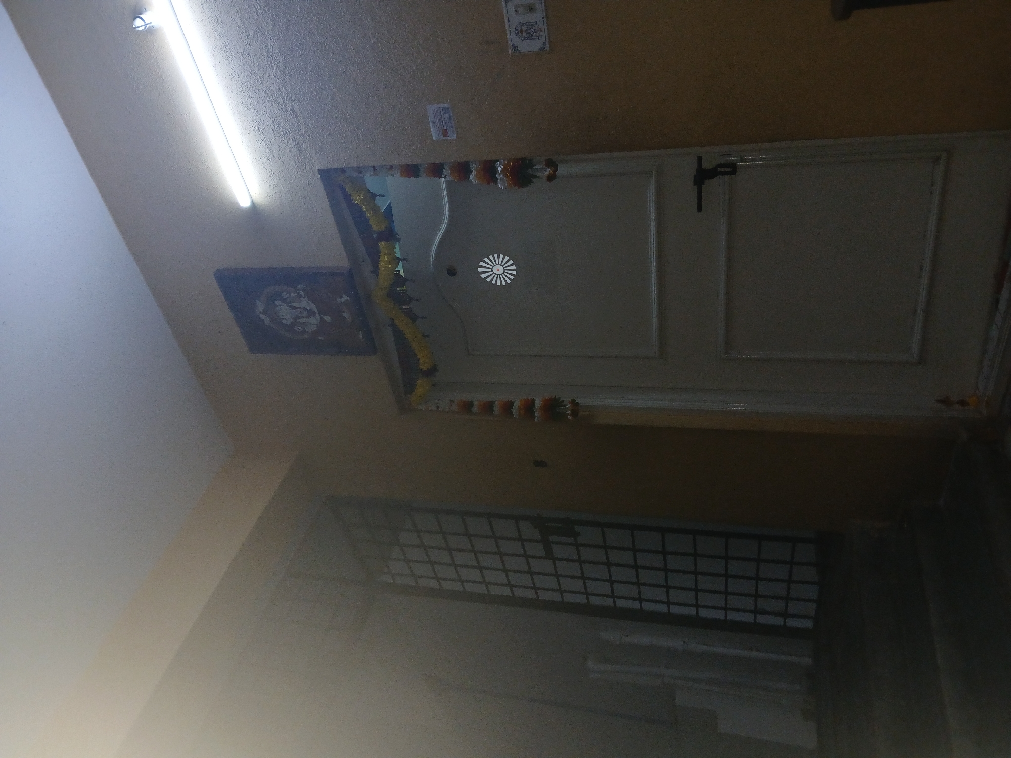 Here's a description of the image:
**Scene:** The image appears to be taken inside a residential building or apartment in Bengaluru, India. The viewpoint is from below, looking upwards towards an interior door.
**Door:** The door is white with a decorative design on the upper panel. It is adorned with garlands of marigolds and other decorative elements, likely for a religious or auspicious occasion.
**Objects:** A religious image hangs to the left of the door. Above the door, there are some small plaques or signs affixed to the wall. Below the door, there appears to be a window or ventilation grill made of metal bars.
**Lighting:** The lighting is provided by a fluorescent tube light near the top of the image.
