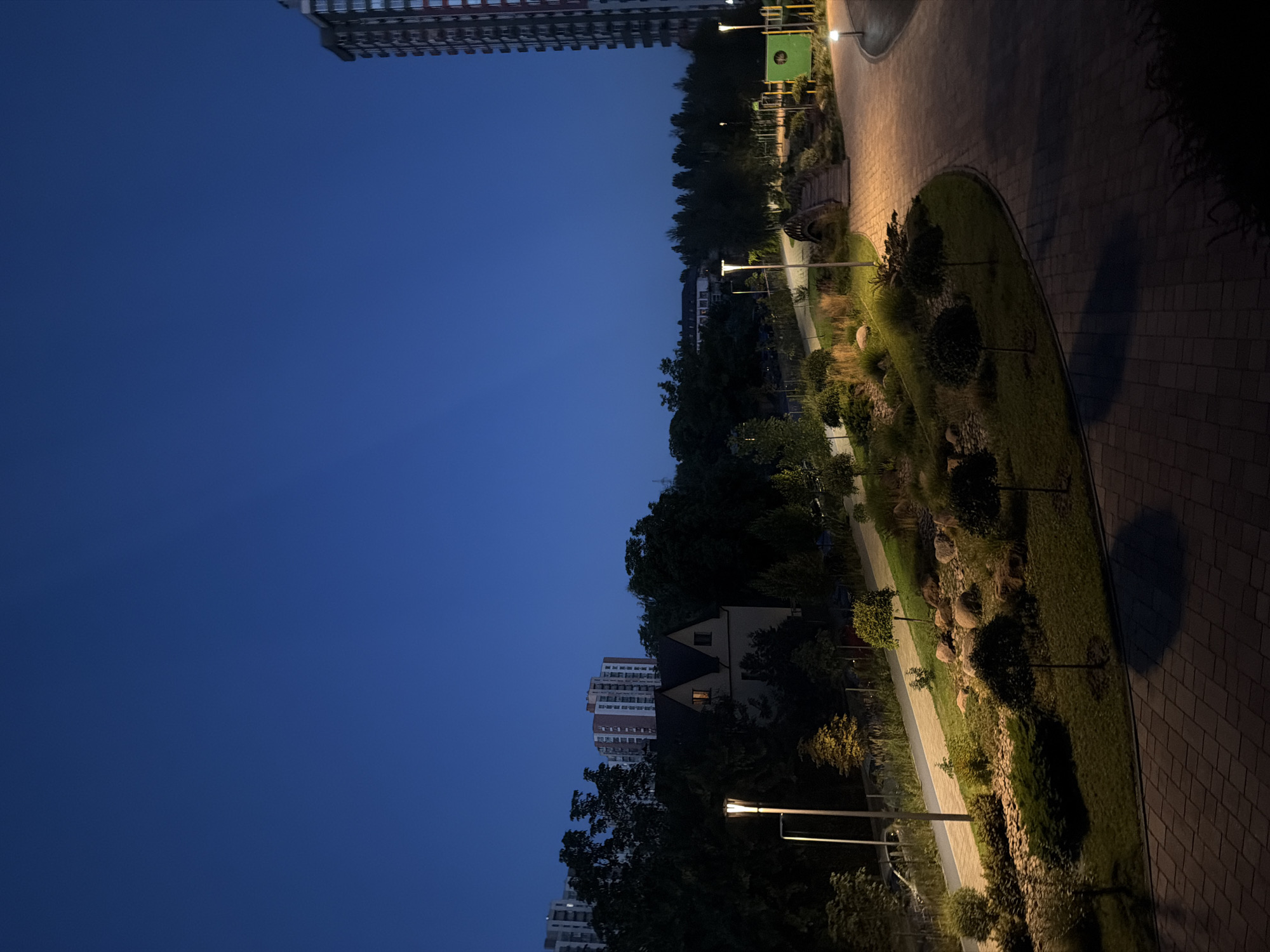 This is a nighttime photograph of a park area in Kyiv, Ukraine. The sky is a deep, dark blue, indicating twilight or early night. In the foreground, a paved pathway with a brick pattern curves through the park. To the left of the path, there is a landscaped area with grass, bushes, and decorative trees, illuminated by a tall lamppost. Further back, a small bridge spans a dry creek bed or rock garden.
In the middle ground, a quaint, gabled house with illuminated windows is nestled amongst dense trees. Behind the house, several multi-story apartment buildings are visible, some with lit windows. The closest apartment building on the right is a modern, tall structure with a visible facade of balconies and windows. There are additional lampposts throughout the park, providing warm, localized lighting. A playground structure with a green element is visible in the distance to the right. The overall scene suggests a residential area with integrated parkland. There are no people visible in the image.