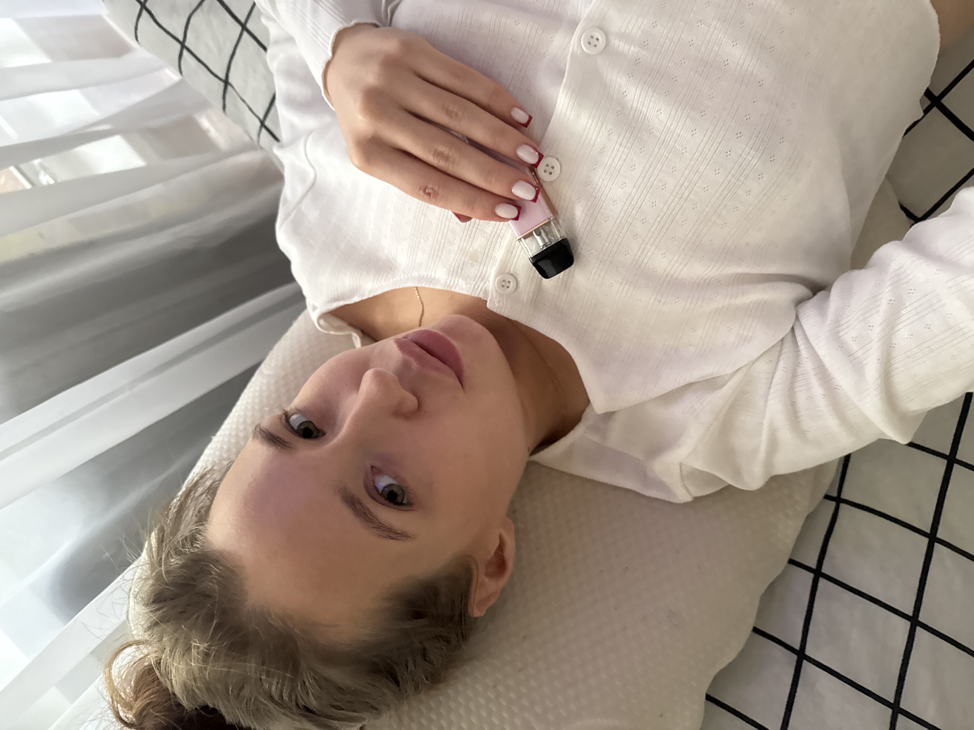 A young woman lies on a bed, looking up at the camera. Her head is on a pillow, and she is wearing a white, button-up cardigan. Her left hand rests on her chest, holding a small, pink and metallic object. She has light brown hair, styled in a messy bun, and her eyes are wide and green. Her lips are slightly parted. She is wearing light makeup, and her fingernails are painted white with a dark red tip. The bed is covered with white bedding that has a black checkered pattern on one side. To the left of the bed, sheer white curtains are visible, suggesting an indoor setting with natural light. The overall mood is relaxed and casual.