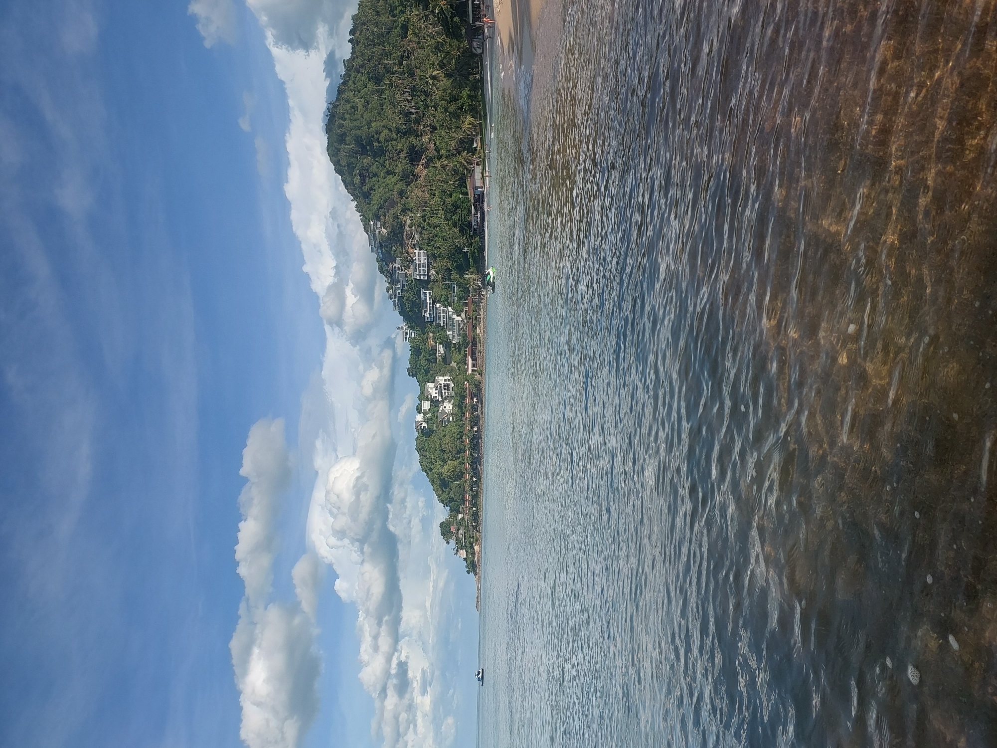 The image depicts a serene coastal scene at Baan Chaweng Noi, Thailand. The foreground is dominated by the calm, clear ocean water, revealing the sandy bottom below. Gentle ripples create a shimmering effect on the surface. In the mid-ground, a sandy beach lines the shore, with scattered palm trees and low-lying buildings nestled among them. The background features a lush green hillside dotted with white houses, ascending towards the top of the frame. A few small boats, including a jet ski, are visible in the water near the shore. The sky is a vibrant blue, adorned with scattered white cumulus clouds, suggesting fair weather. The overall atmosphere is one of tranquility and natural beauty.