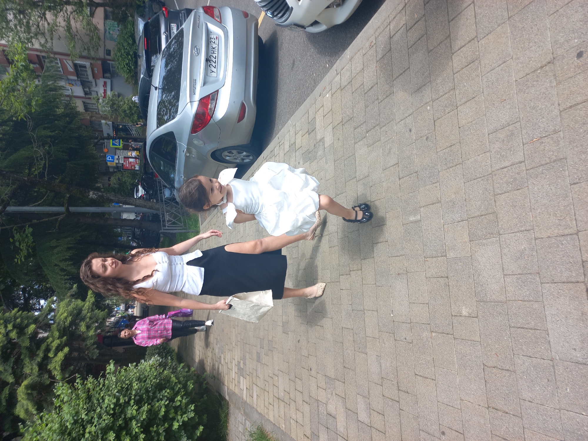 The image shows a woman and a young girl in Sochi, Russia. The woman, with long curly brown hair, is wearing a white top and a black skirt. She is carrying a white shopping bag. The young girl is wearing a white dress and black shoes. The girl is standing with one leg extended, seemingly dancing. They are walking on a paved sidewalk next to a street where several cars are parked. A silver Hyundai with a license plate "Y222HK23" is visible. Another person in a pink plaid shirt is walking further behind them. Trees and buildings are visible in the background.