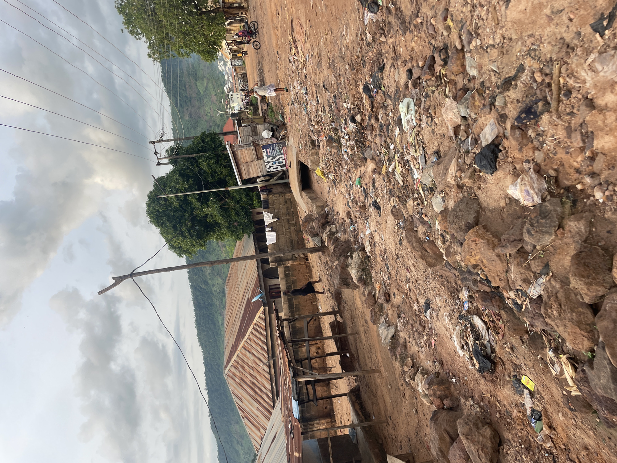 The image depicts a scene in Lokoja, Nigeria, characterized by a dirt road that is heavily littered with trash, including plastic bags, wrappers, and other debris. The road is flanked by low-lying buildings with corrugated iron roofs, some appearing weathered. A few people are visible: one person in a black dress walks down the road, another person in white shorts walks near a small wooden kiosk labeled "POS" and "Offset Ventures," and further down the road, a person on a motorcycle is present. Trees and hills form the backdrop under a cloudy sky. Power lines are strung across the scene. The overall impression is one of a somewhat underdeveloped or neglected urban environment.