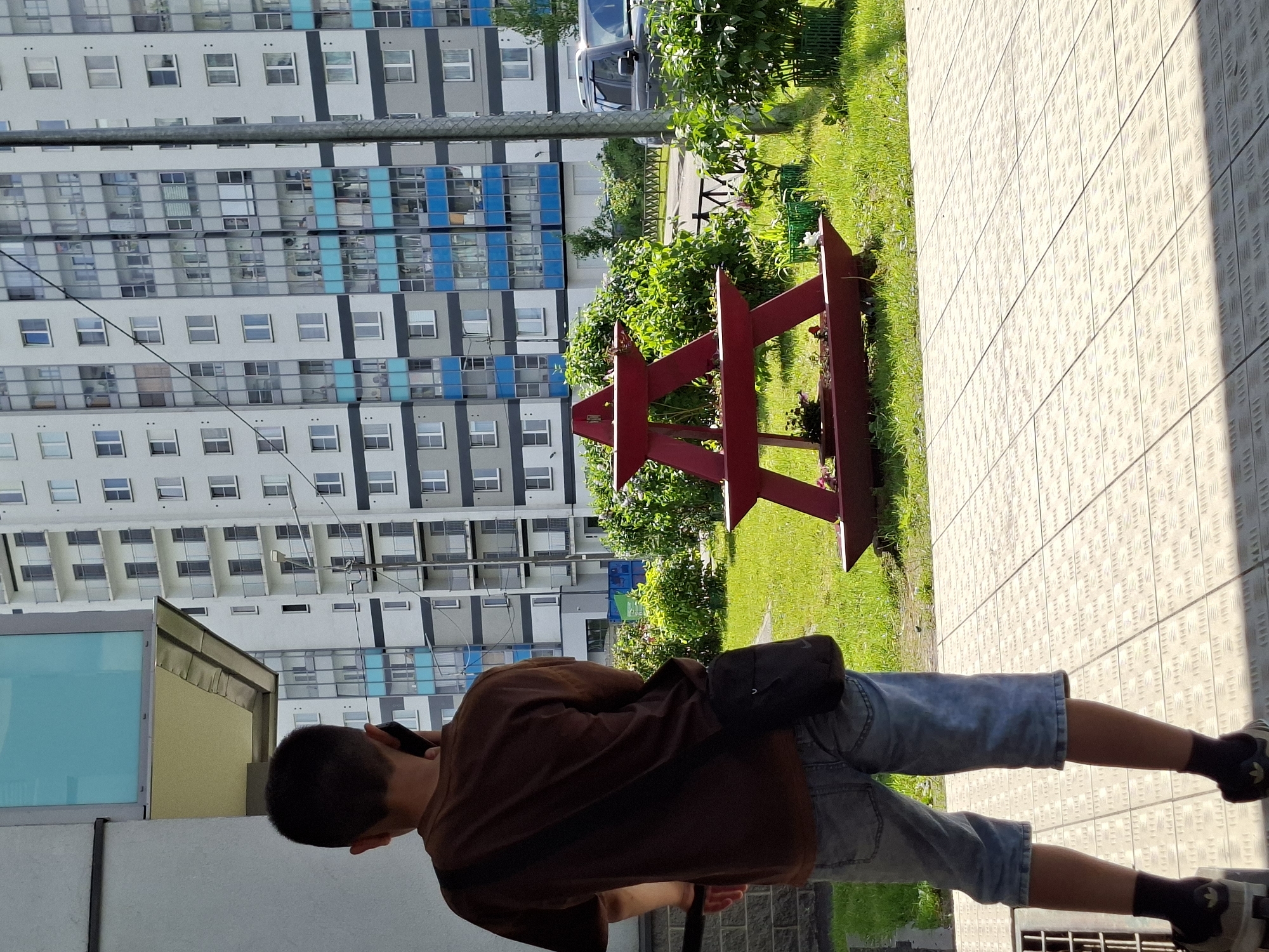 An outdoor scene in what appears to be a residential area in Saint Petersburg, Russia, is captured in bright daylight. The image features a person, seen from the back, standing on a patterned paved surface. The individual is wearing a brown t-shirt, light blue denim shorts, dark socks, and dark shoes, and has a black shoulder bag. They are holding a smartphone to their ear, suggesting they are engaged in a phone call.
To the right of the person, a red wooden decorative structure with angled, possibly star-like, elements stands on a patch of green grass. Beyond the grass and bushes, a paved road is visible, with a small, light-colored car partially seen in the background. Dominating the left side of the frame is a tall, modern residential building with numerous windows, balconies, and distinctive blue and grey accent panels on its facade. Some reflections are visible in the building's windows, and power lines or cables cross in front of it at mid-height. The ground surface where the person stands is a light-colored, textured pavement, resembling a diamond plate or checker plate pattern. The clear, bright lighting indicates a sunny day, casting distinct shadows.