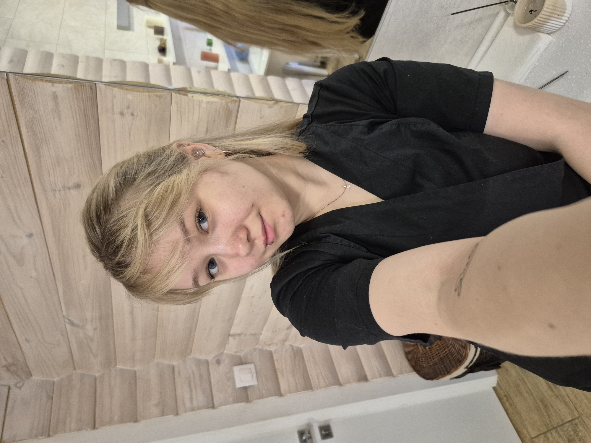 A fair-skinned person with blonde hair and blue eyes is taking a selfie. The person is wearing a black t-shirt and has a lip piercing. Behind the person is a wall with a herringbone pattern. In the upper-left corner, there is a reflection of another person with long blonde hair. A dark wicker basket is partially visible in the lower right corner. The location cannot be ascertained with the provided image.