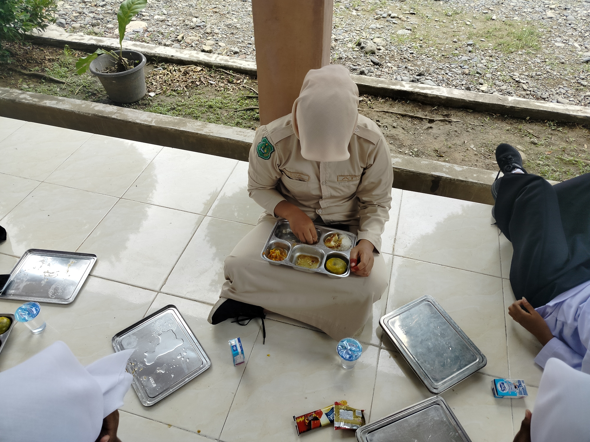 The image depicts an outdoor scene in Lawebekung, Indonesia, where three individuals are gathered on a tiled patio. The central figure, wearing a beige uniform and headscarf, is seated and eating from a compartmentalized meal tray. To their left and right, other individuals are partially visible, one of whom is wearing a white head covering and another in a black garment. Several metal trays, a small carton of milk, a cup of water, and some snack wrappers are scattered on the ground around them, indicating a mealtime or break. The background reveals a stony, unpaved area with scattered vegetation and a potted plant, suggesting a simple outdoor setting, possibly within a school or community compound. The overall atmosphere appears casual and informal.