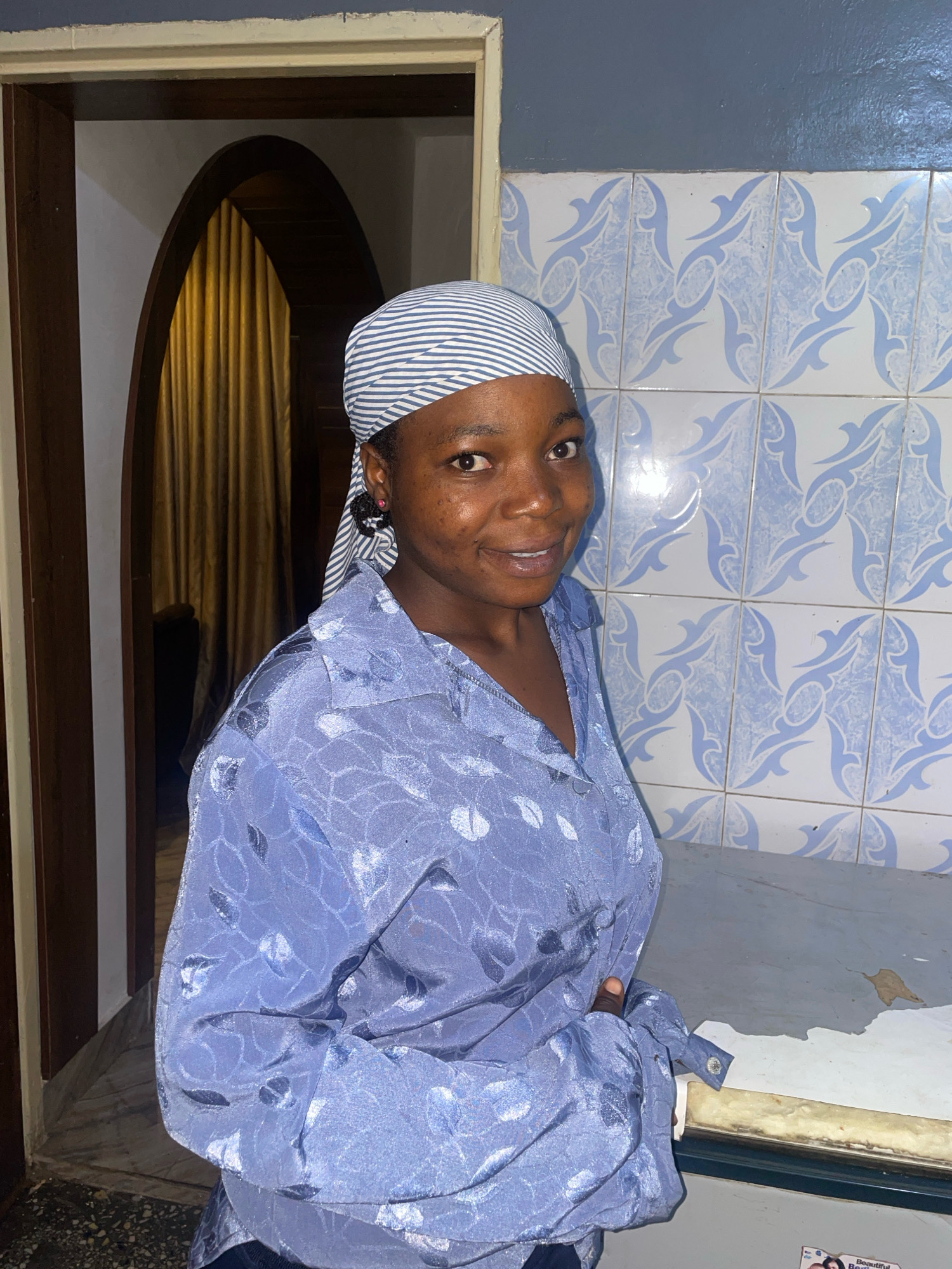A young woman wearing a blue and white striped headscarf and a patterned blue shirt stands in front of a tiled wall. She is smiling at the camera. Behind her, an arched doorway leads to a room with golden curtains. The scene appears to be an interior domestic setting, possibly in Jos, Nigeria.