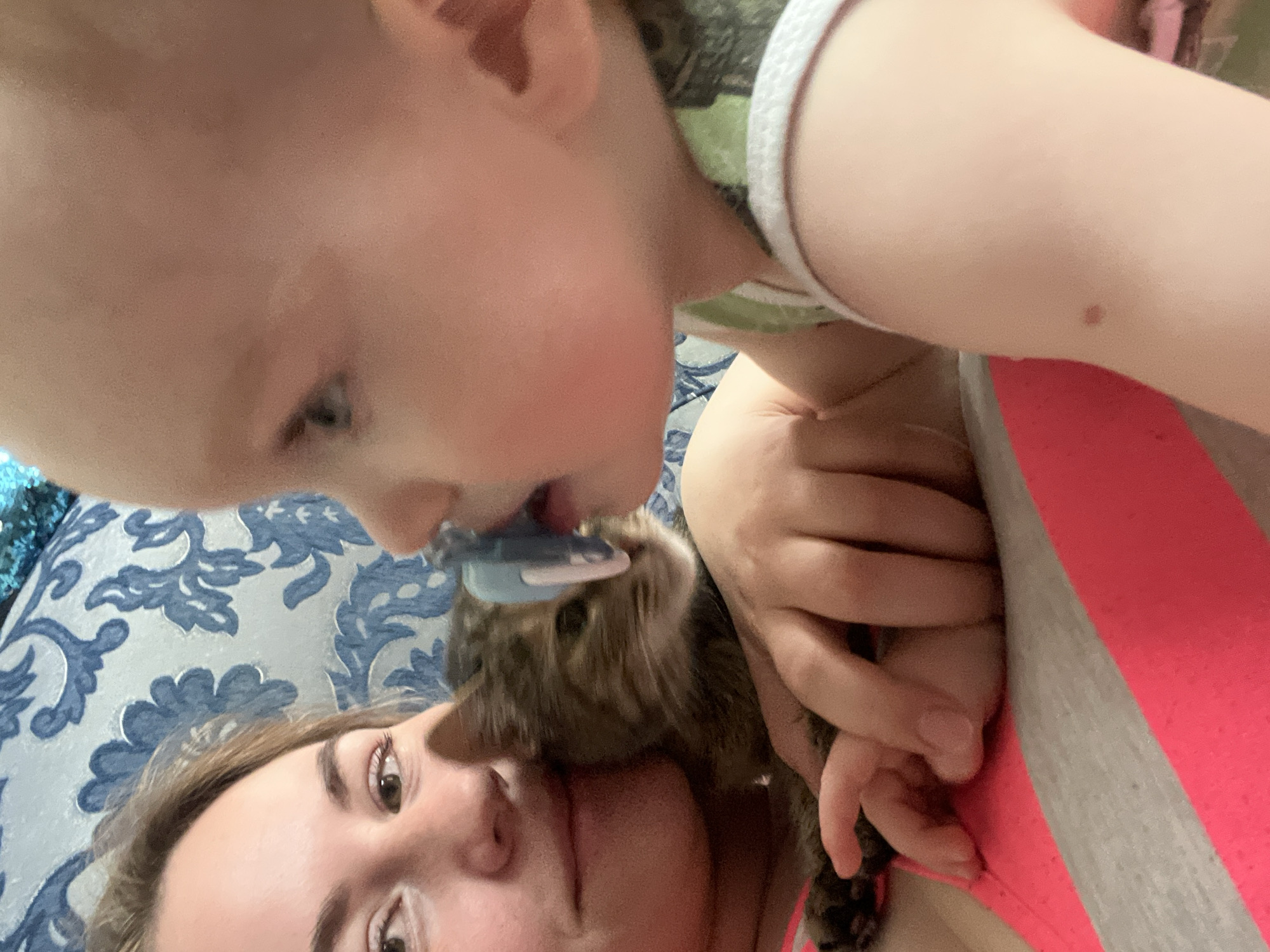 The image shows a close-up, angled shot of a woman lying on a blue and white patterned surface. A child with a pacifier is positioned above her, with a cat nestled between them. The woman is looking at the camera, while the child appears to be interacting with the cat. The scene is indoors. The location of the image is unknown, and thus cannot be placed in the city of Chaikovsky, Russia, or any other location.