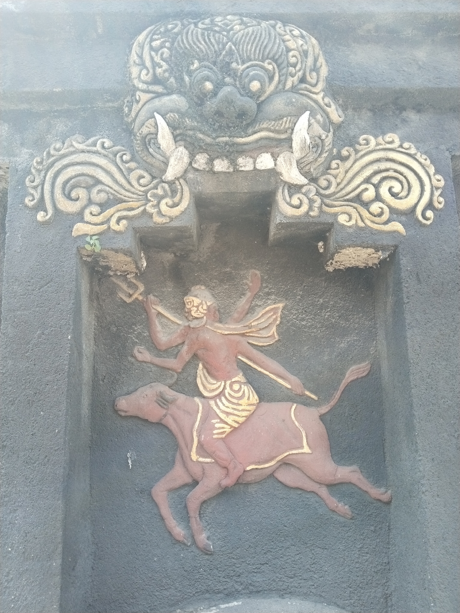A close-up shot depicts a stone carving, likely part of a temple or similar structure, located in Masaran, Indonesia. The carving features a multi-armed figure riding a bull. The figure has a reddish-brown complexion and is adorned with gold-colored patterns around the waist. One hand holds a trident, while another extends outward. Above the figure and the bull is a decorative motif of a monster-like face with prominent teeth and swirling patterns. The background is a dark gray, textured surface, suggesting the material of the wall or structure. The overall image conveys a sense of ancient artistry and cultural significance.