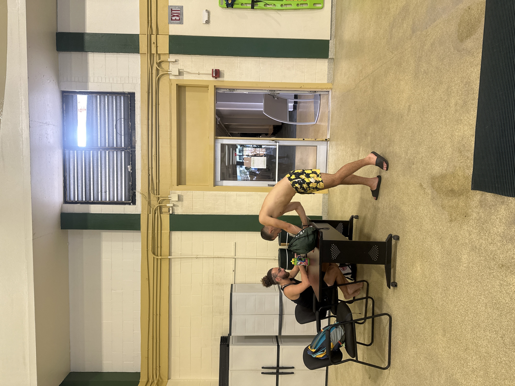 The image, taken in an indoor setting likely a swimming pool or recreational facility in Tampa, USA, features two men engaged in casual activities. The walls are tiled with a combination of white and cream, accented by a dark green horizontal stripe, while the floor is a light tan or beige.
One man, shirtless and clad in yellow and black patterned swim trunks and black flip-flops, stands in the mid-ground. He is leaning over a black metal table, seemingly rummaging through a dark green bag placed on it.
The second man, sporting curly hair and a beard, is seated at the same table, wearing a black tank top and shorts, and is barefoot. He is drinking from a colorful bottle. Behind him, several white plastic storage units are stacked against the wall.
The background includes a window with vertical slats to the left, through which bright daylight shines, indicating it is daytime. To the right, there's a wider opening, possibly a serving hatch or reception window, alongside a doorway. Above this doorway, a red "EXIT" sign is visible. A green spineboard or rescue board is mounted on the wall near the exit. Beige conduits and pipes run along a wall pillar. A dark, ribbed mat lies on the floor to the far right of the frame.