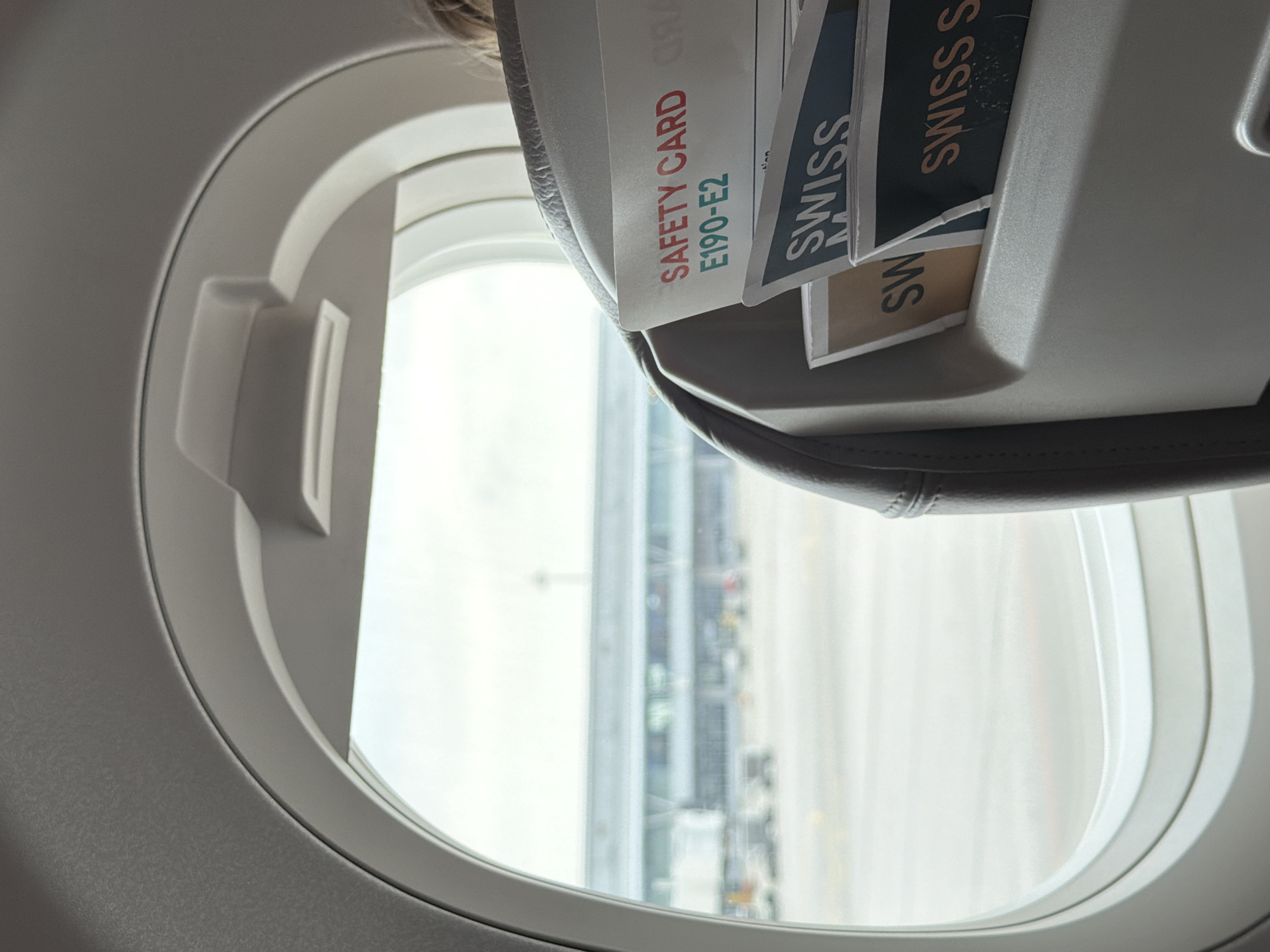 The view from an airplane window shows an airport terminal with several Swiss International Air Lines safety cards and brochures. The safety card reads "SAFETY CARD E190-E2". The location cannot be determined to be Sofia, Bulgaria from the provided image.