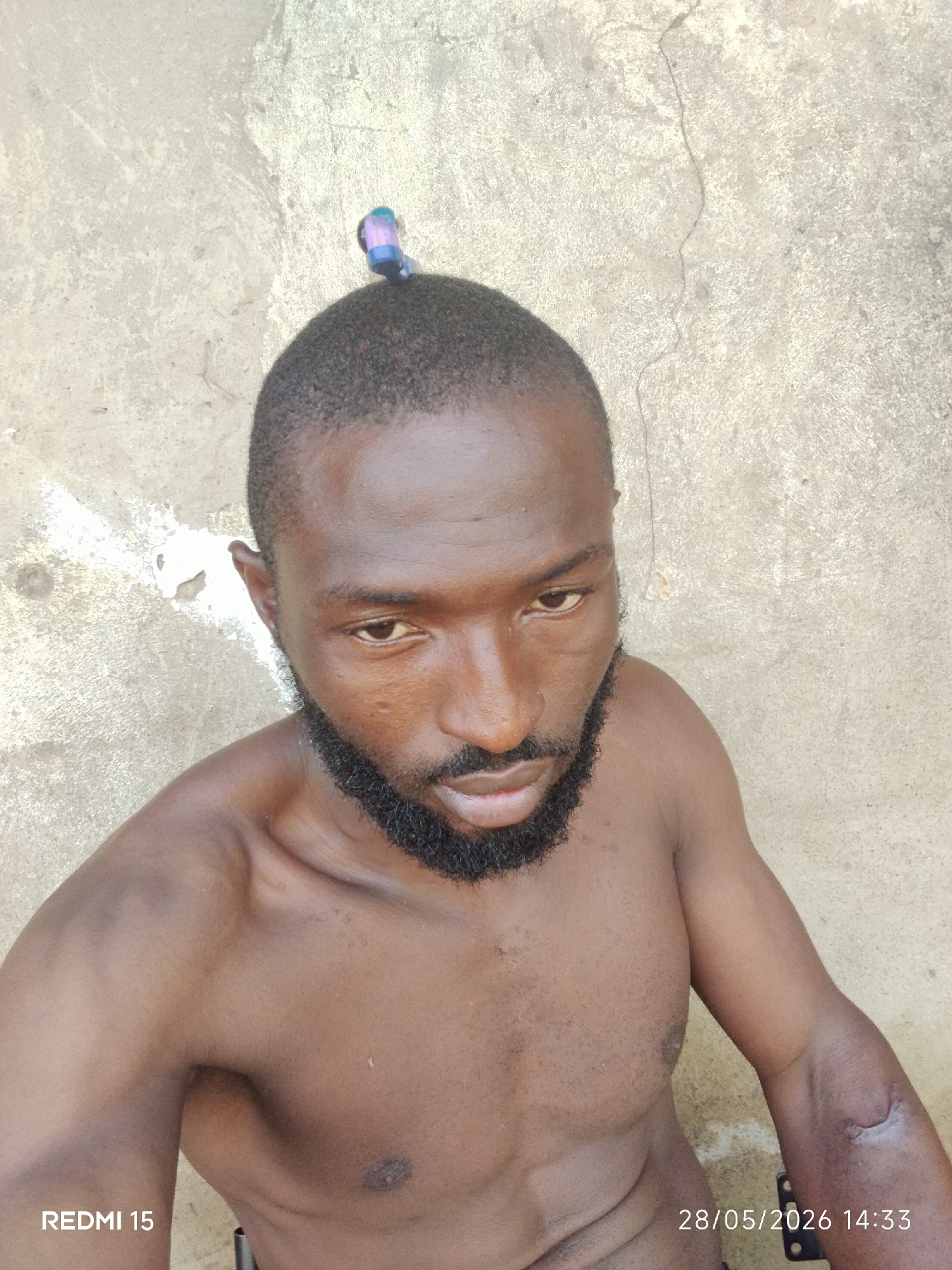 A medium shot shows a shirtless, dark-skinned man with a closely shaved head, a full black beard, and a small blue and pink object clipped to the top of his head. He is looking down at the camera. The background is a beige concrete wall with patches of discoloration. The words "REDMI 15" appear in the lower-left corner, and "28/05/2026 14:33" are in the lower-right corner. No specific indications point to Sabon Tasha, Nigeria; the location is inferred from possible metadata.