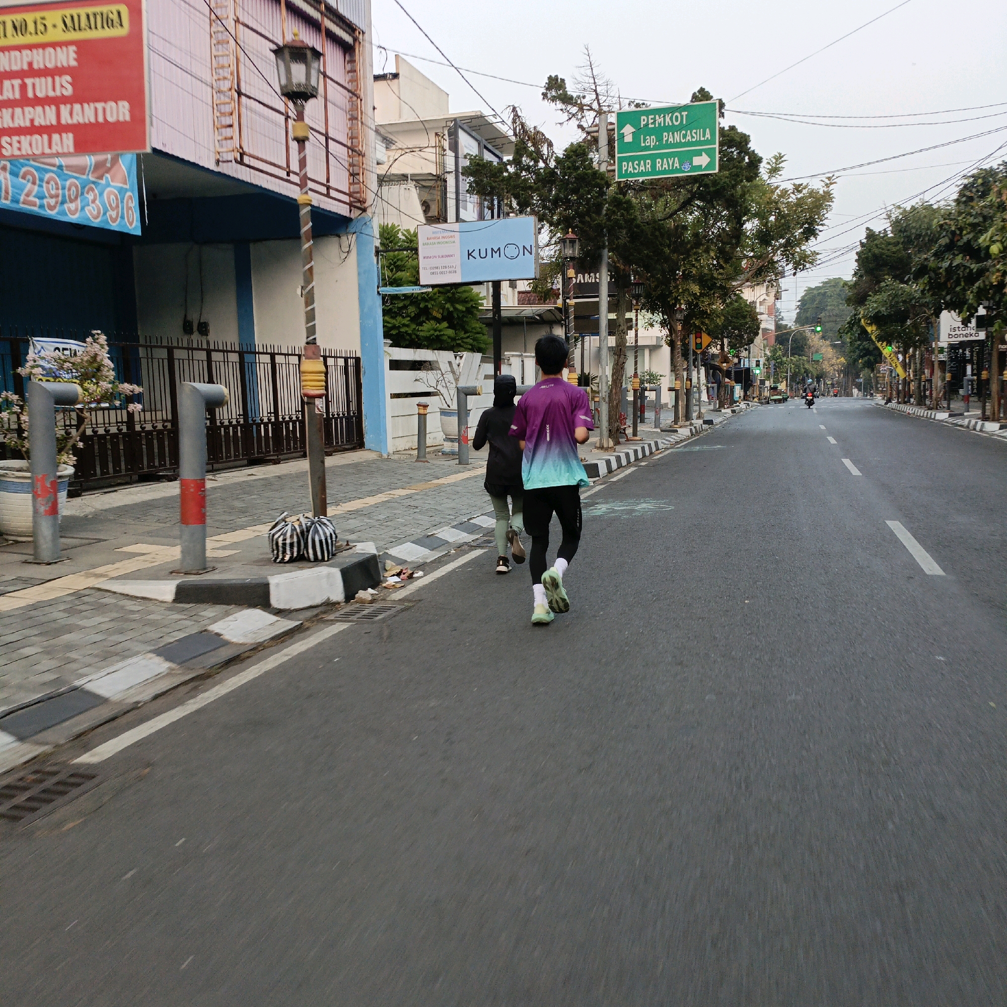 Two individuals are running on the asphalt road of Salatiga, Indonesia. The person in front is wearing a purple and teal gradient t-shirt and black leggings, while the person behind them is wearing a black hoodie and light green leggings. The road has a white dashed line dividing the lanes, and a sidewalk with black and white striped curbing is visible to the left. On the sidewalk, there are two grey cylindrical bollards with red accents and two striped bags. A street lamp with a golden base and decorative elements stands near the sidewalk. In the background, buildings with signs and trees line the street. A green directional sign indicates "PEMKOT, Lap. PANCASILA" to the left and "PASAR RAYA" to the right. The sky is overcast.