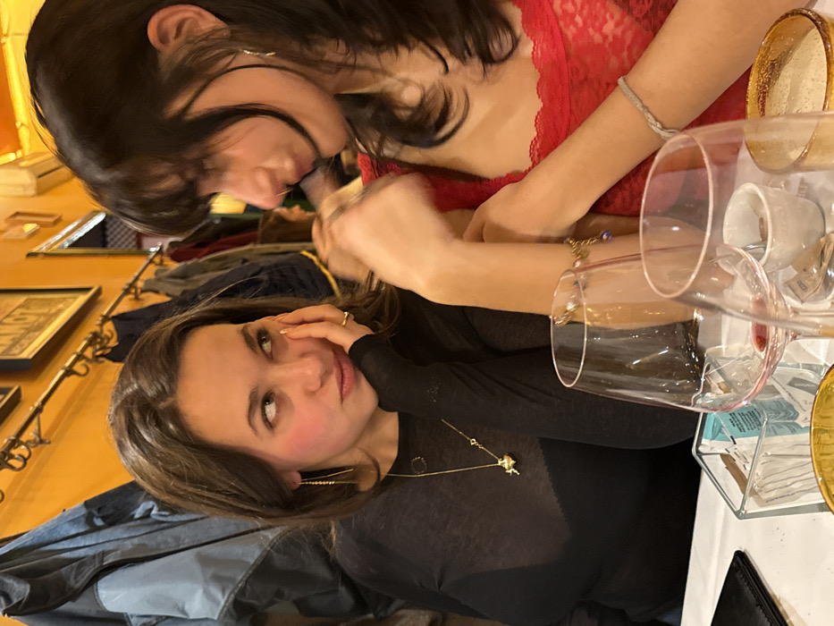 Two young women are at a table. One is wearing a black mesh top and gold jewelry, looking at the camera with a slight smile. The other is in a red lace top, with dark hair and bangs, leaning towards the first. Wine glasses and a gold charger plate are on the table. Coats hang on a wall in the background, along with framed artwork. The scene is lit with a warm, indoor light.