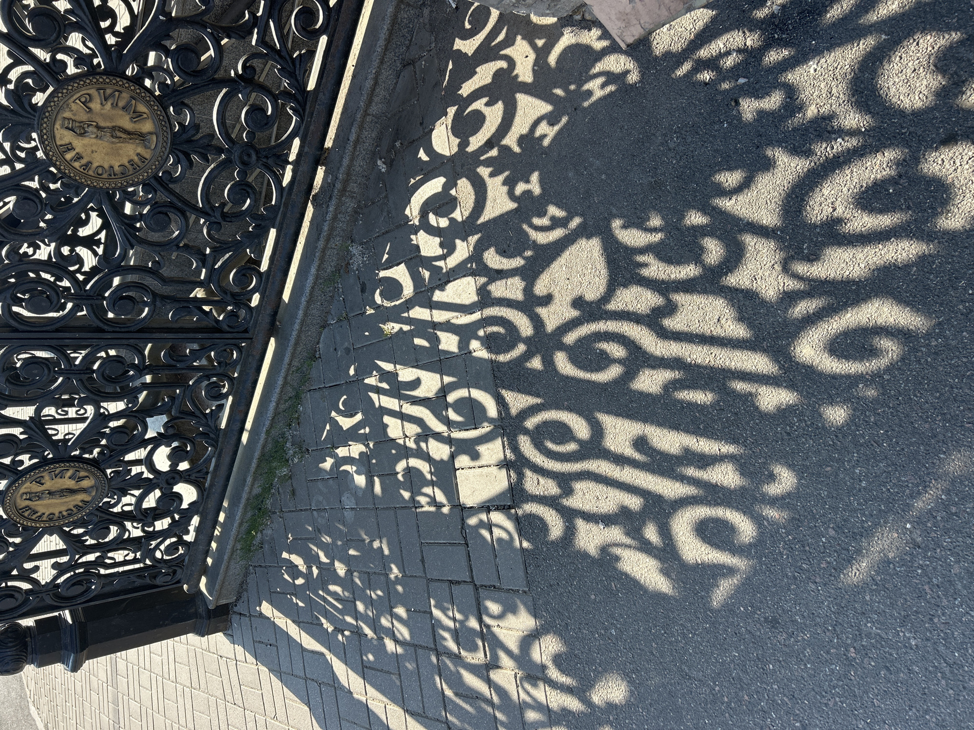 The image shows the decorative shadow of an ornate black metal gate cast on a paved surface and asphalt. The gate features intricate scrollwork and two circular medallions. One medallion is visible at the top left, and another is partially visible at the bottom left. The medallions appear to be made of a golden-colored metal and contain embossed text and imagery. The text on the visible medallion reads "РИМ" at the top and "РЕСТОРАН" along the bottom curve. The medallion also contains an image of a reclining figure.
The shadow is sharply defined, suggesting a bright, sunny day. The light is coming from an angle, indicating it is likely morning or late afternoon. The shadow covers both the textured paving stones and the darker asphalt, highlighting the complex pattern of the gate's design. The setting appears to be outdoors, possibly on a street or sidewalk next to a building. The overall mood is tranquil, with the interplay of light and shadow creating an artistic and visually appealing composition. There are no people visible in the image. The ground is clean, with no visible debris. The colors are dominated by the dark metal of the gate, the golden medallions, the grey paving stones, and the dark asphalt, all illuminated by the bright sunlight.