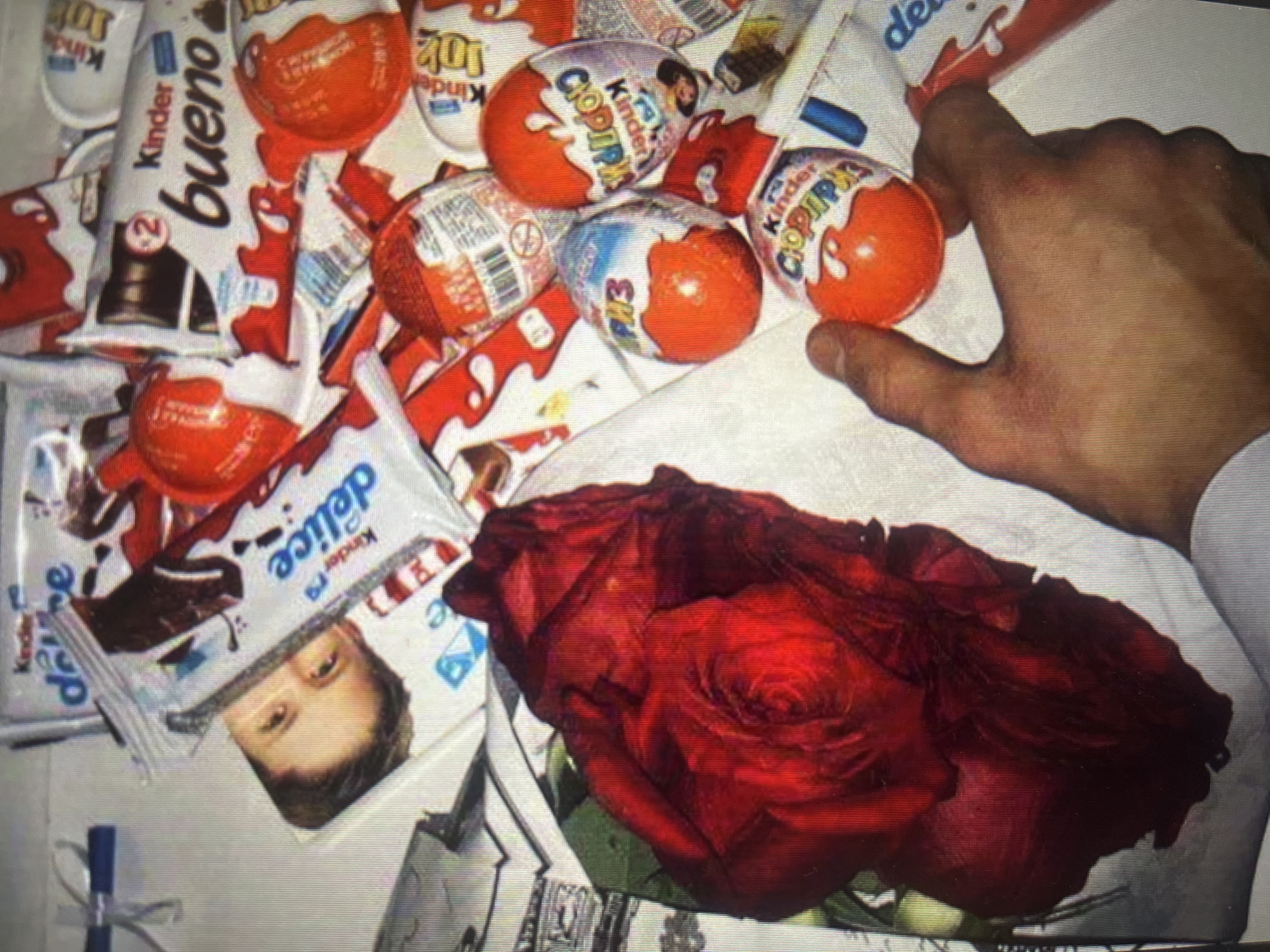 This is a close-up, high-angle, eye-level shot of a collection of Kinder brand chocolates and a red rose, all arranged on a white surface, likely a table or bed. The products are scattered across the frame, with some overlap. Prominent among the items are several Kinder Surprise eggs, identifiable by their orange plastic shells and multicolored wrappers. There are also several bars of Kinder chocolate, including "Kinder Bueno" and "Kinder Delice." A single, large, dark red rose is positioned in the lower left quadrant of the image, its petals unfurled. In the bottom right corner, a person's hand is visible, holding one of the Kinder Surprise eggs. The overall lighting appears to be artificial and diffused. The scene does not provide any specific visual cues to indicate its location within the city of Moscow, Russia.