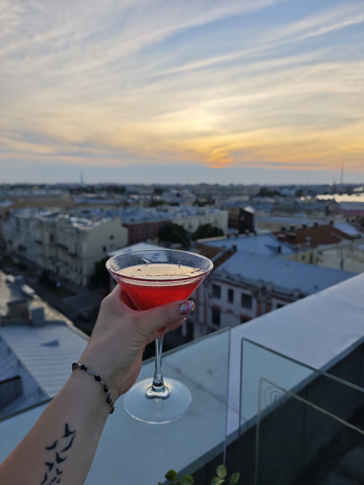 A hand holds a martini glass filled with a red cocktail against the backdrop of a city skyline during sunset. The glass is a clear martini glass with a wide rim, containing a vibrant red liquid. The hand has a bracelet on the wrist and a tattoo of birds in flight on the forearm. The city in the background appears to be Saint Petersburg, Russia, with its characteristic architecture, including buildings with tiled roofs and historical facades. The sky displays a gradient of colors, from pale blue at the top to warm oranges and yellows near the horizon, indicating the sun is setting. A modern glass barrier is visible in the foreground, suggesting the scene is from a rooftop bar or terrace.