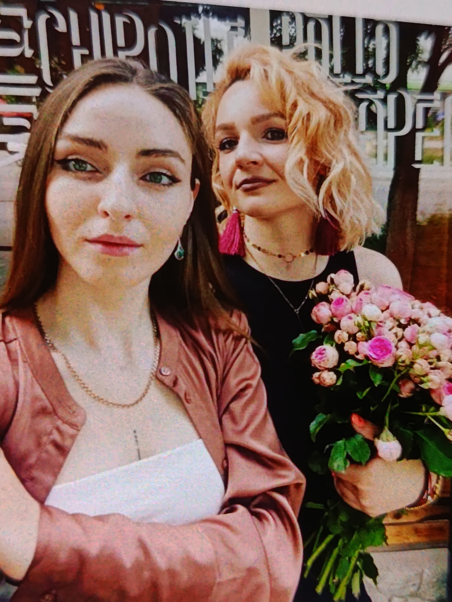 Two young women are posing for a selfie in front of a building with large, stylized white lettering that reads "CHIROPRACTIC BALTI" on a dark background. The woman on the left has long, dark brown hair and is wearing a light pink satin cardigan over a white top. She has green eyes and is looking directly at the camera. The woman on the right has blonde, curly hair and is wearing a black dress. She has large, pink tassel earrings and a gold necklace. She is holding a bouquet of pink roses and baby's breath. The background appears to be an outdoor setting, possibly a street or a park, with trees visible behind the building. The image was taken in the city of Bălți, Moldova.