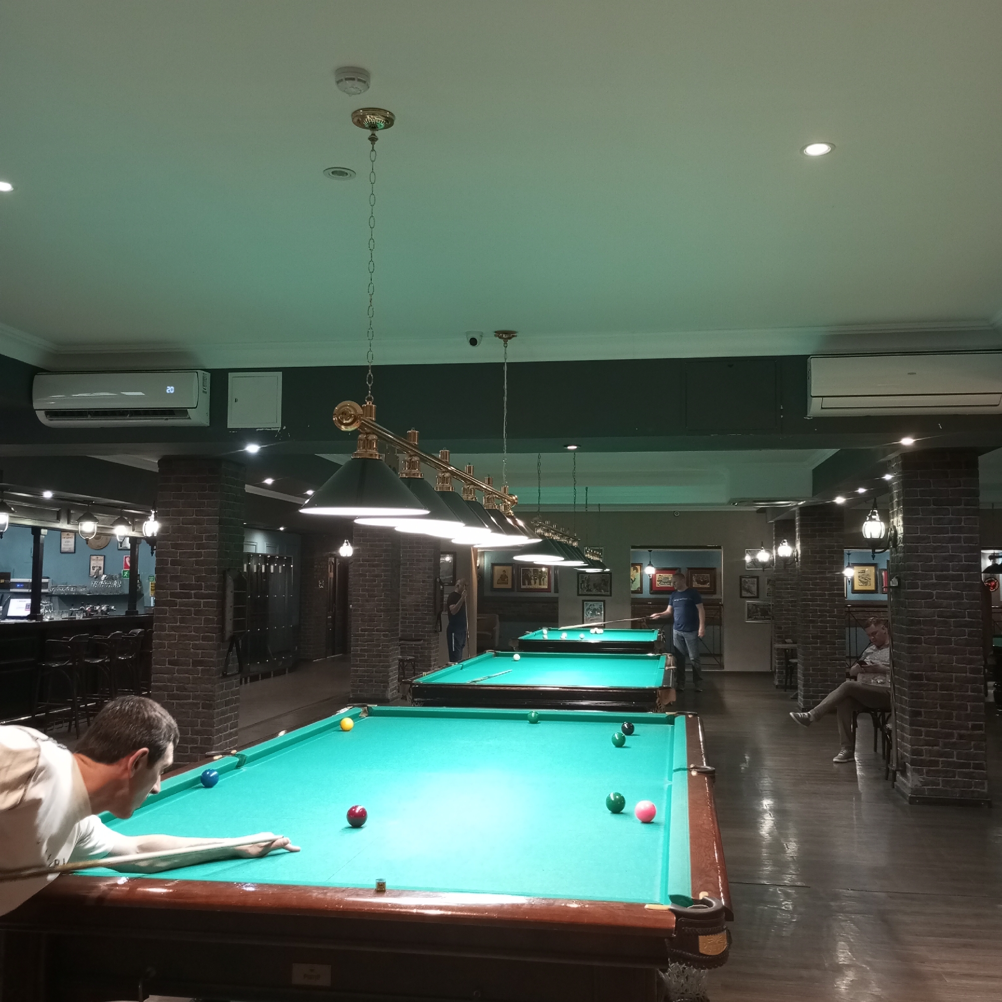 This is an indoor photograph of a billiard hall. The room is illuminated by overhead lighting and has multiple billiard tables. In the foreground, a man is bent over a billiard table, preparing to strike a ball with his cue. Another man is standing near a billiard table further back in the room, holding a cue. A third man is seated on a chair, looking at his phone. The hall has brick-textured walls and a polished wooden floor. The lighting fixtures are ornate, hanging from the ceiling. In the background, there appears to be a bar area with seating. The overall atmosphere is that of a recreational establishment. The image appears to have been taken in Izhevsk, Russia.