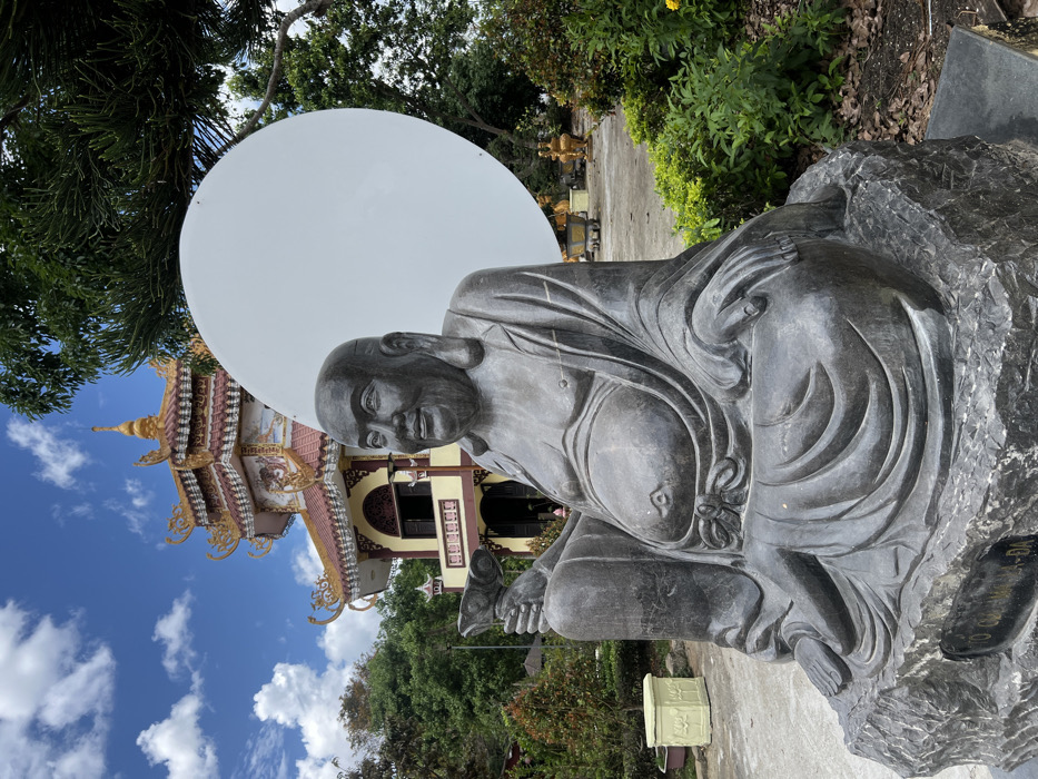 A dark grey stone statue of Budai, commonly known as the Laughing Buddha, sits prominently in the foreground, facing slightly right with a smiling expression and a visible belly. The statue holds a pot or bowl in its raised left hand, while its right hand rests on its knee. It is mounted on a rough-hewn dark grey stone base, which displays a carved inscription including "NAM MO CHUONG LAI HA-DA".
Behind the statue, a multi-tiered Buddhist pagoda rises under a clear blue sky with scattered white clouds. The pagoda features light yellow walls, dark brown trim, and red-tiled roofs adorned with golden decorative elements and finials. Windows are visible on the upper tier. To the right and behind the statue's head, a large, plain white circular disc stands vertically. The surrounding area comprises green foliage, including bushes, trees, and a light yellow planter. Further in the background, smaller golden statues are discernible on the temple grounds. The scene is located in the city of Đăk Mar, Vietnam.