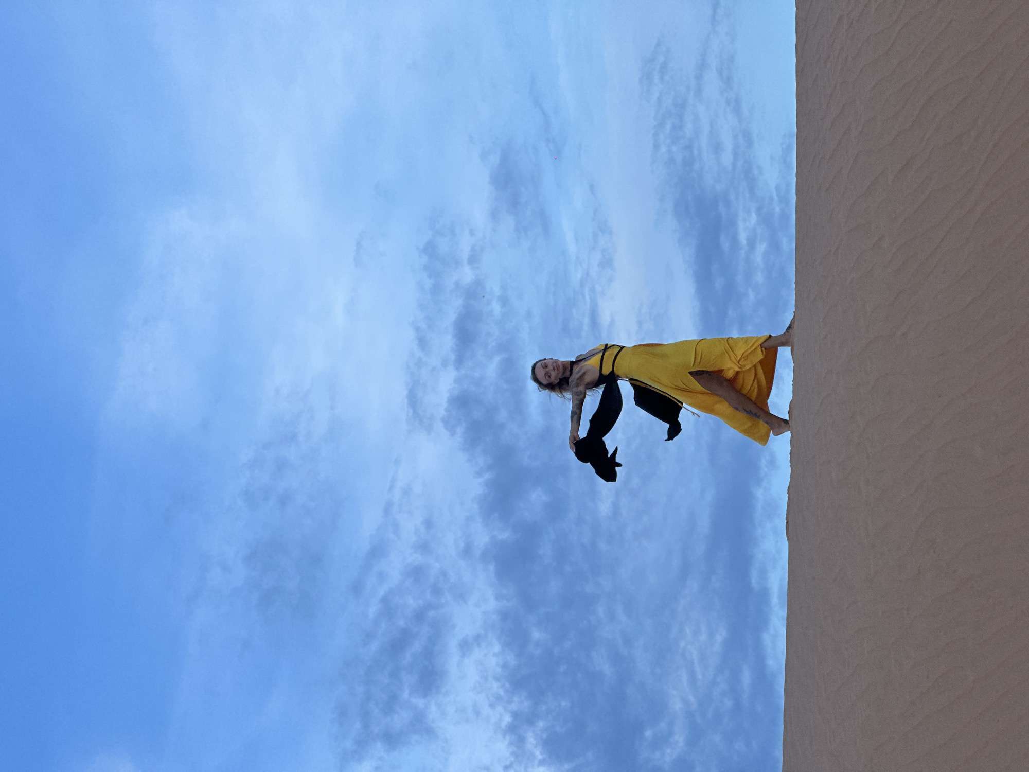 A person is captured mid-air, appearing to dance or leap in a dramatic pose against a backdrop of a cloudy blue sky and a sand dune. The person is wearing a bright yellow dress with black straps and black pants. Their body is arched backwards, with one leg extended. Their arms are also extended, and they are holding a black piece of fabric. The image is oriented such that the sand dune is on the right side and the sky fills the left. The sun is not directly visible, but the bright sky suggests it is daytime. The weather appears to be clear with scattered clouds, and the scene is set in a sandy environment, likely a desert or beach, consistent with the location context of Mui Ne, Vietnam. The pose and attire suggest a moment of artistic expression, perhaps dance or performance. No text is visible in the image.