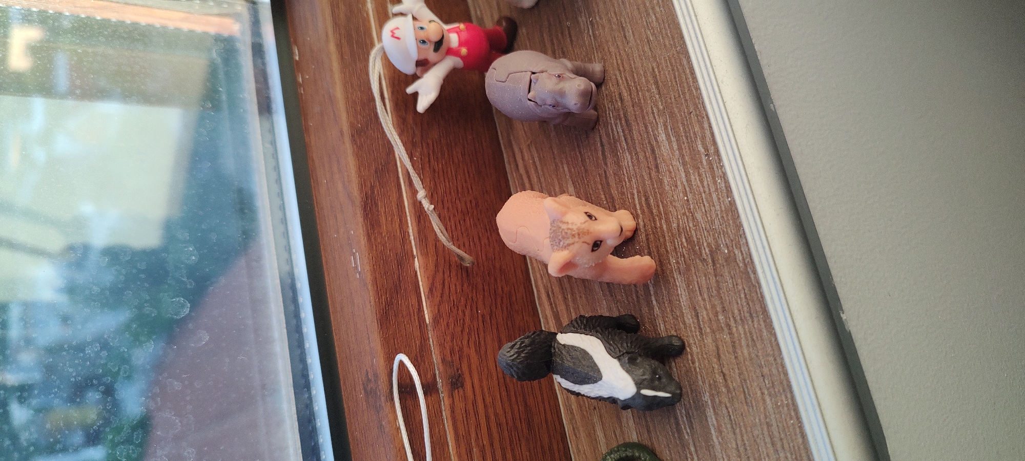 The image shows a windowsill, likely inside a home, featuring an assortment of small toy figures. The toys include a figure resembling the Mario character, a grey hippopotamus puzzle piece, a pig puzzle piece, and a skunk figure. The windowsill has a wood-grain finish. A window with a blurry view is on the left, and a portion of a white wall is visible on the right. The location is unknown.