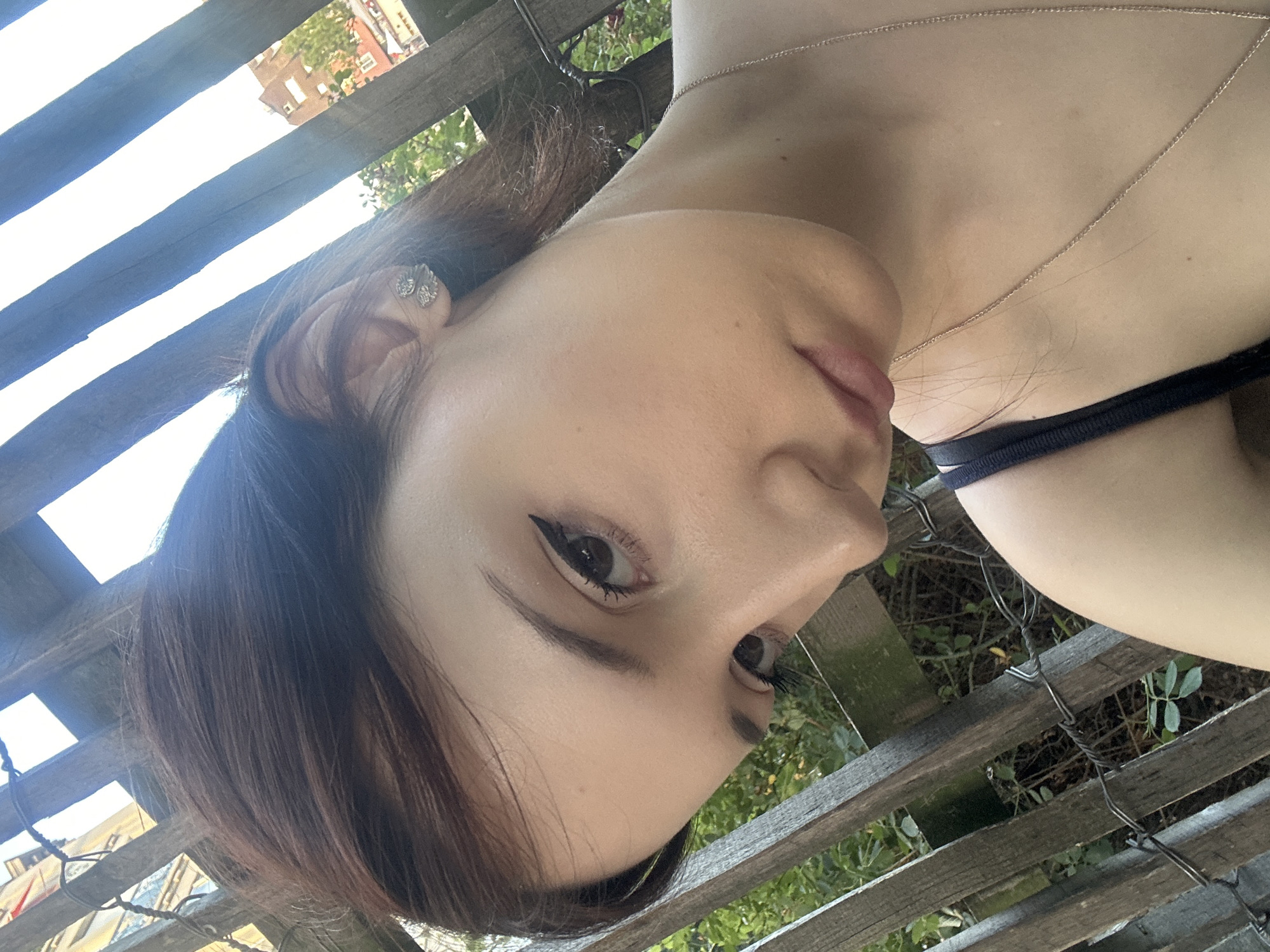 The media file shows a close-up, rotated view of a person's face and upper body. The person has dark hair, fair skin, and is wearing eye makeup. They are also wearing a delicate silver necklace and a dark-colored garment that appears to be a bra strap. The background suggests an outdoor setting with wooden fencing and greenery. The sunlight suggests it is daytime. In the distance, a building with red-tiled roofs is visible, hinting at a European architectural style. The image seems to capture a candid moment, possibly taken outdoors.