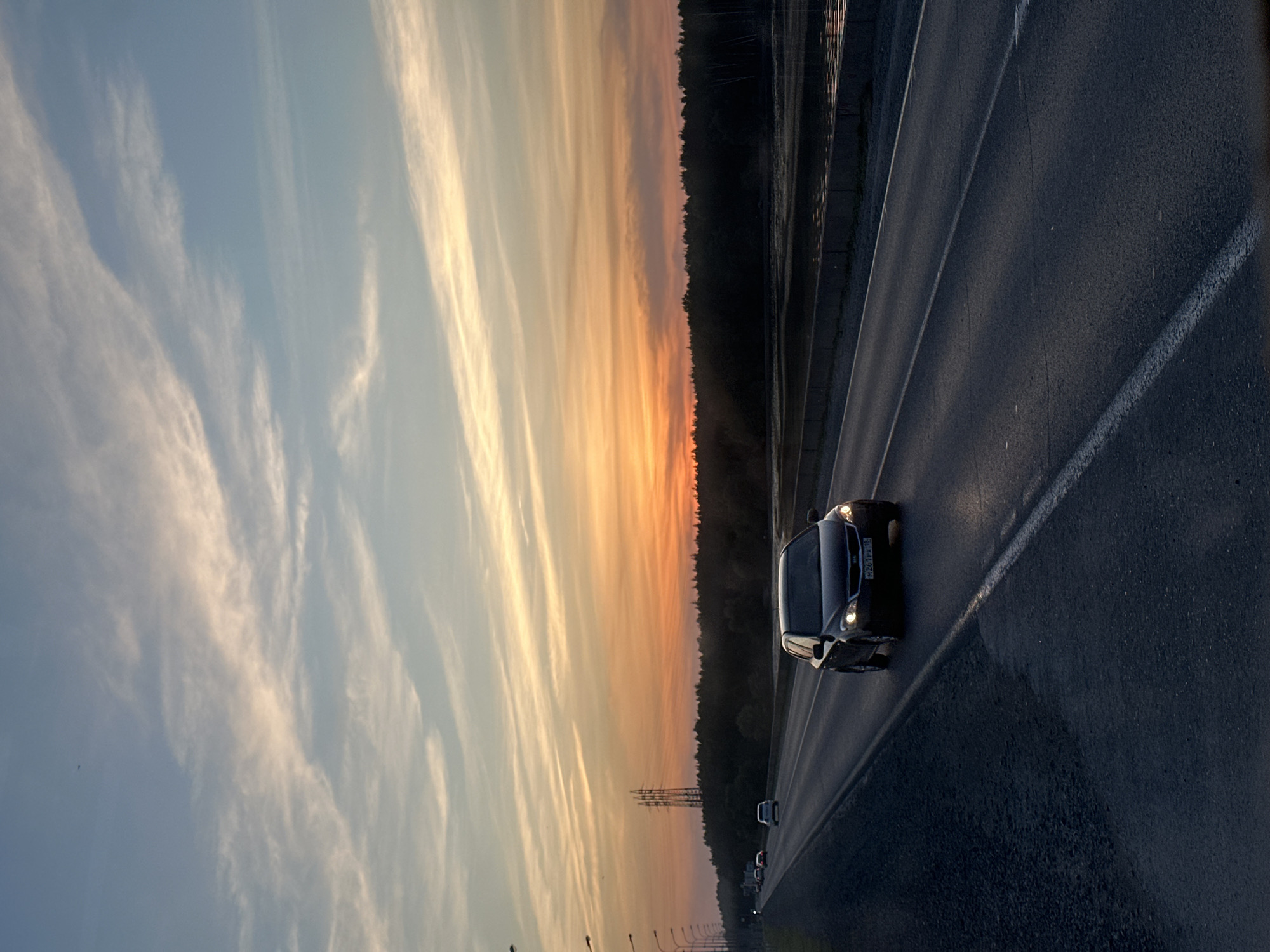 A slightly distorted, rotated, eye-level shot captures a car speeding along a highway at sunset, near the city of Noviy, Russia. The sky transitions from a light blue with wispy clouds in the upper left, to orange hues near the horizon. Alongside the road, a line of dense trees creates a dark vertical border. The pavement is primarily dark and textured, with white lines marking the lanes. The car, positioned centrally on the road, has its headlights illuminated, and its license plate reads "P224TP178". Other vehicles can be seen further down the highway. A series of power pylons are visible in the distance near the horizon.