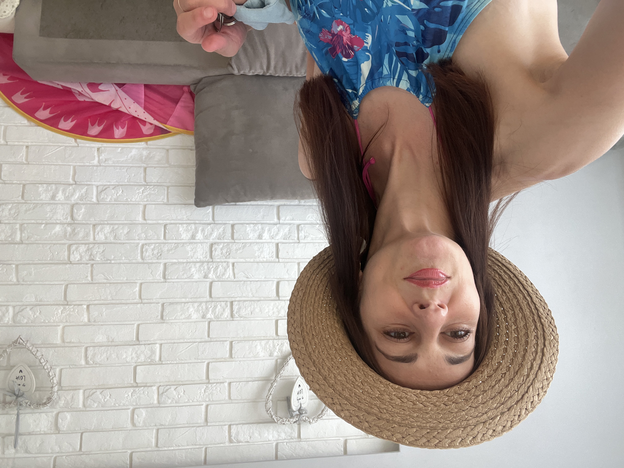 This is an upside-down selfie of a woman wearing a straw hat and a blue swimsuit with a floral pattern. She has long, dark brown hair and is wearing makeup, including pink lipstick. In the background, there is a white brick wall with a heart-shaped decoration that says "Love". To the left of the woman, part of a gray sofa and a pink and white patterned mat are visible. The image appears to have been taken indoors, likely in a home environment, possibly before or after a beach outing given the attire. The lighting suggests it could be daytime. The woman's expression is neutral.