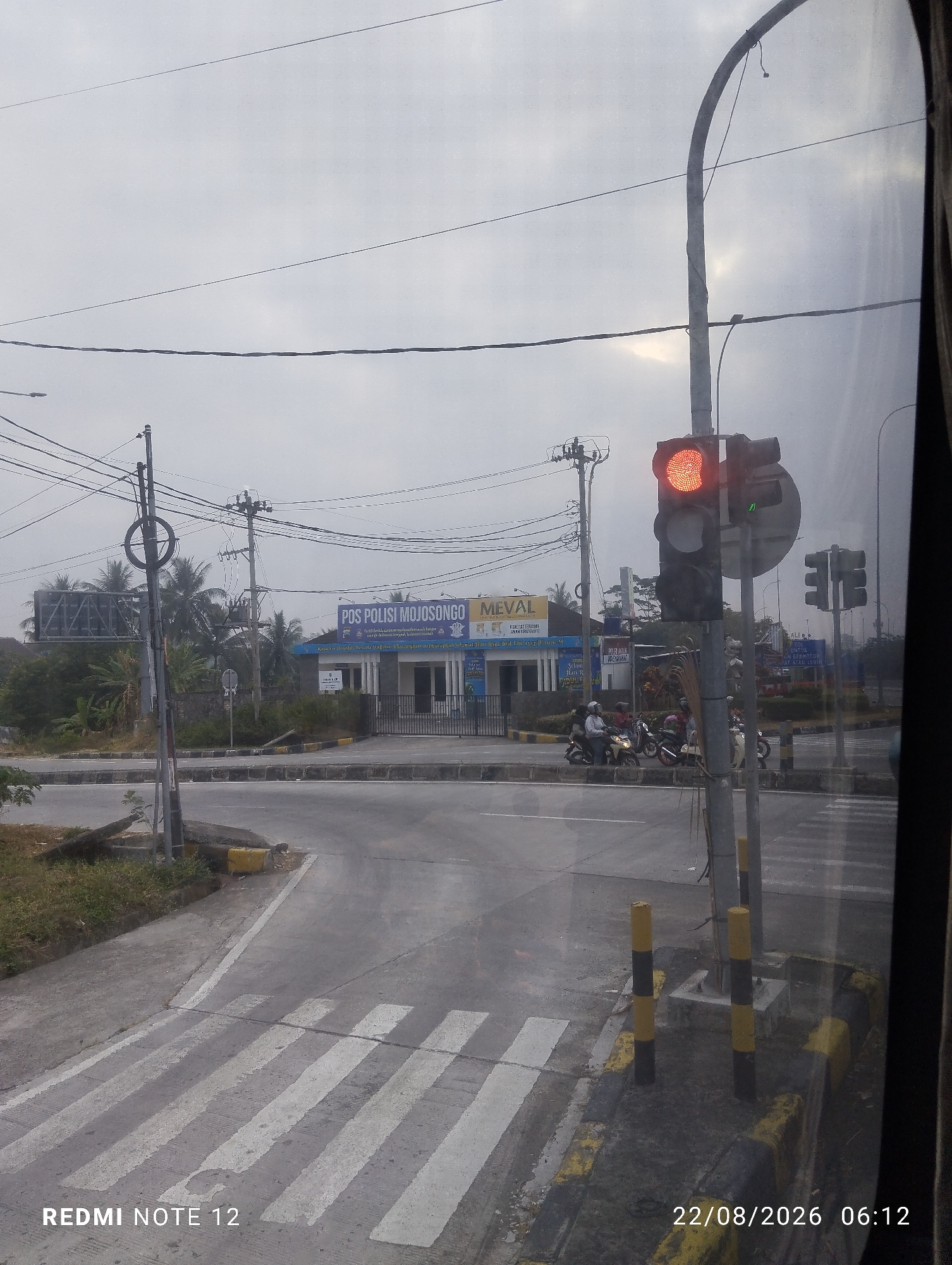 The image shows a street scene at an intersection in Dipan, Indonesia, under an overcast sky. A traffic light is visible and illuminated red, indicating a stop. Several motorcycles are waiting at the intersection. To the left, a building with signage reads "POS POLISI MOJOSONGO" and "MEVAL," suggesting a police post and a business. Overhead, power lines crisscross the sky, supported by utility poles. A pedestrian crossing is marked on the road. The lower right corner of the image shows a timestamp: 22/08/2026 06:12, and the text "REDMI NOTE 12" in the lower left corner. The environment appears to be a semi-urban area with some vegetation and buildings.