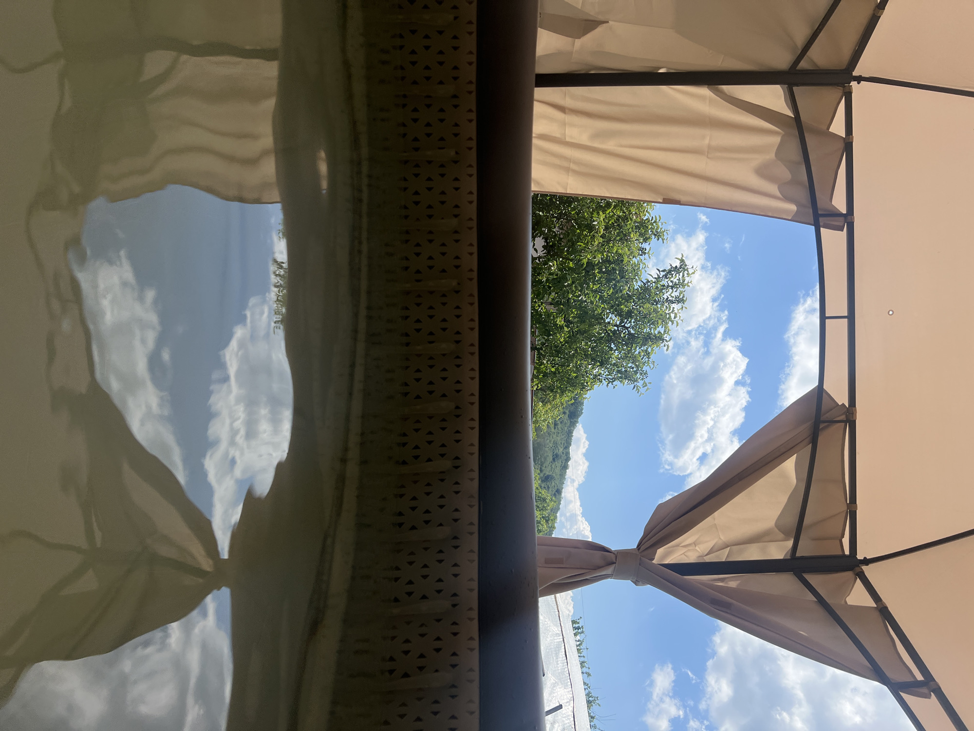 The image shows a view looking upwards from underneath a beige gazebo structure. Metal supports and fabric curtains are visible, framing a view of a blue sky with white clouds and a green tree-covered hill. The leftmost part of the image shows a reflective surface, possibly water, mirroring the sky and clouds above. The location appears to be outdoors, potentially in a garden or patio setting. Without additional information or context, it is impossible to confirm that the image was taken in Gorno Kamartsi, Bulgaria.