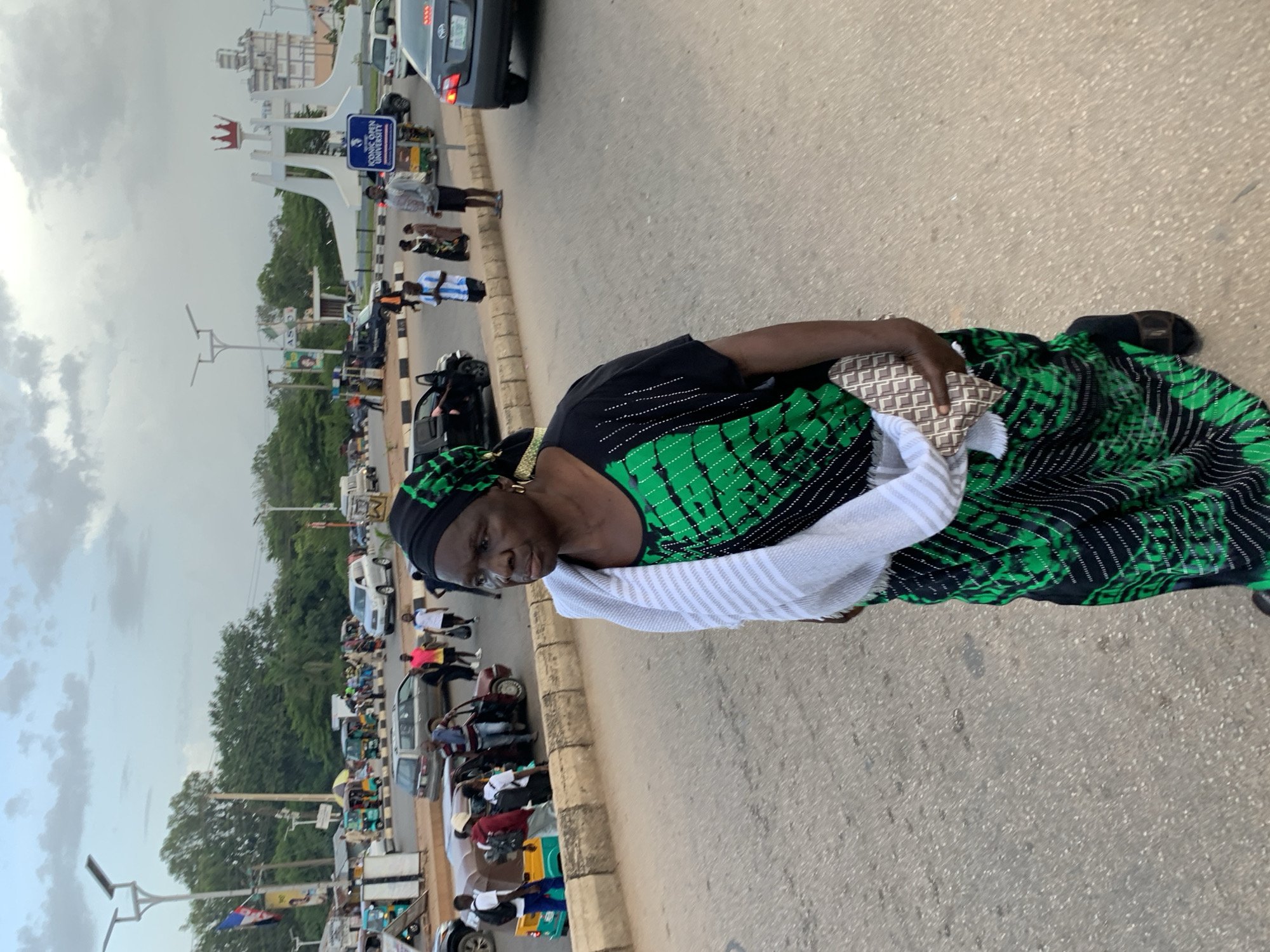 An outdoor, eye-level, medium shot shows a Black woman in the foreground of a street scene in Jos, Nigeria. The woman, who is looking to her left, is wearing a black and green patterned dress, a black headscarf with green accents, a white scarf draped over her shoulders, and dark sandals. She is carrying a patterned clutch bag in her right hand. In the background, a roundabout features a large, white, abstract sculpture with a crown-like top and a blue sign reading "ICONIC OPEN UNIVERSITY". Multiple vehicles, including cars, SUVs, and auto-rickshaws, are visible on the road, along with other pedestrians. The sky is cloudy.