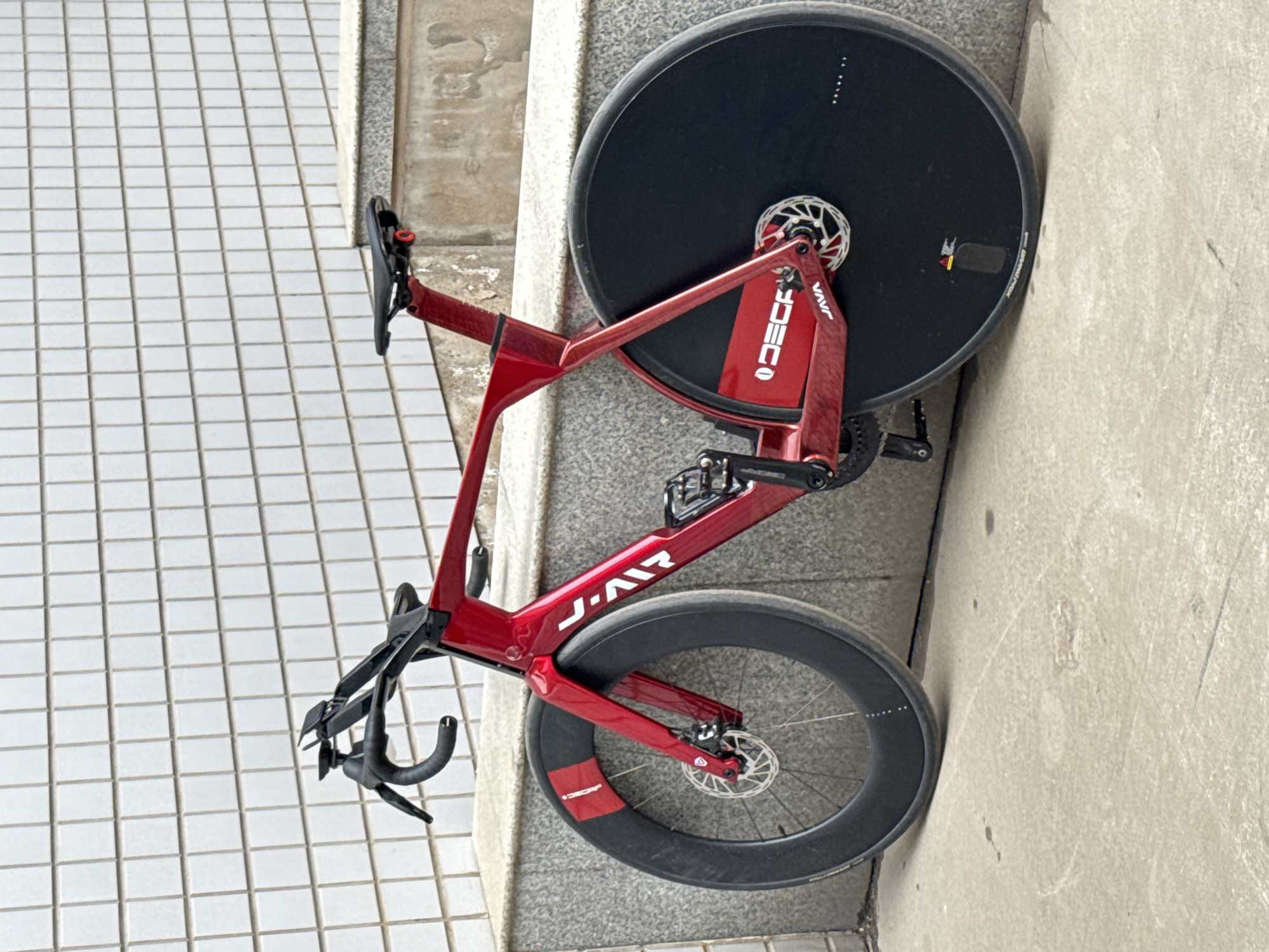 A red Java J-Air time trial bicycle is leaning against a concrete curb separating a tiled sidewalk from a concrete surface. The bicycle has disc brakes and deep profile wheels. The rear wheel is a disc wheel. The bicycle has a water bottle cage mounted on the frame. The location is Zhongtun, China. No people are visible.