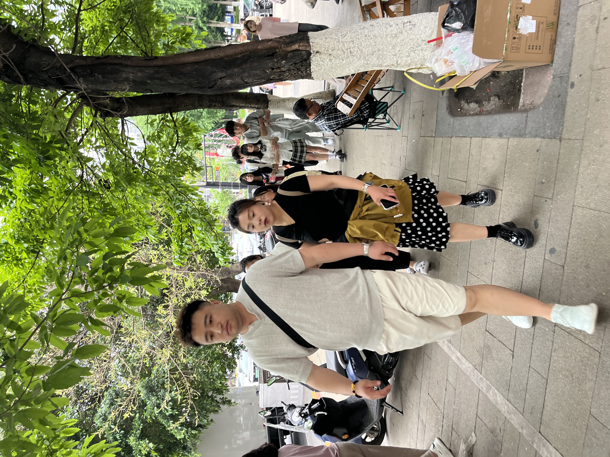 This media file captures an active daytime street scene in Yuzhong District, China. The image is taken from a slightly elevated perspective, looking down a paved sidewalk lined with trees and various individuals.
In the immediate foreground, a man in a light grey short-sleeved shirt and cream-colored shorts stands next to a dark-colored scooter or motorcycle. A black strap from a bag crosses his chest. Behind him, a woman wearing a black top, a mustard-yellow crossbody bag, and a black polka-dot skirt holds a smartphone in her left hand. She is wearing thick-soled black shoes.
Further back along the sidewalk, a group of people is visible. One person is seated on a folding chair next to a tree, playing a wooden percussion instrument that resembles a xylophone. Near them, a small group of individuals, some appearing to be students in white shirts and plaid skirts, stand together. Another person in a pink shirt is seen further down the path near a tree with a white-painted base.
The environment is distinctly urban, characterized by a grey paved sidewalk and mature trees with lush green foliage providing overhead shade. Some tree bases are painted white. To the right of the frame, a large cardboard box filled with plastic bags and what appears to be discarded items rests on the ground. Faint text and an "UP" arrow are visible on the side of the cardboard box. Several scooters or motorcycles are parked along the edge of the sidewalk. The lighting suggests it is daytime, with an even, possibly overcast, light. The overall mood is casual, with people going about their daily activities in this bustling street setting.